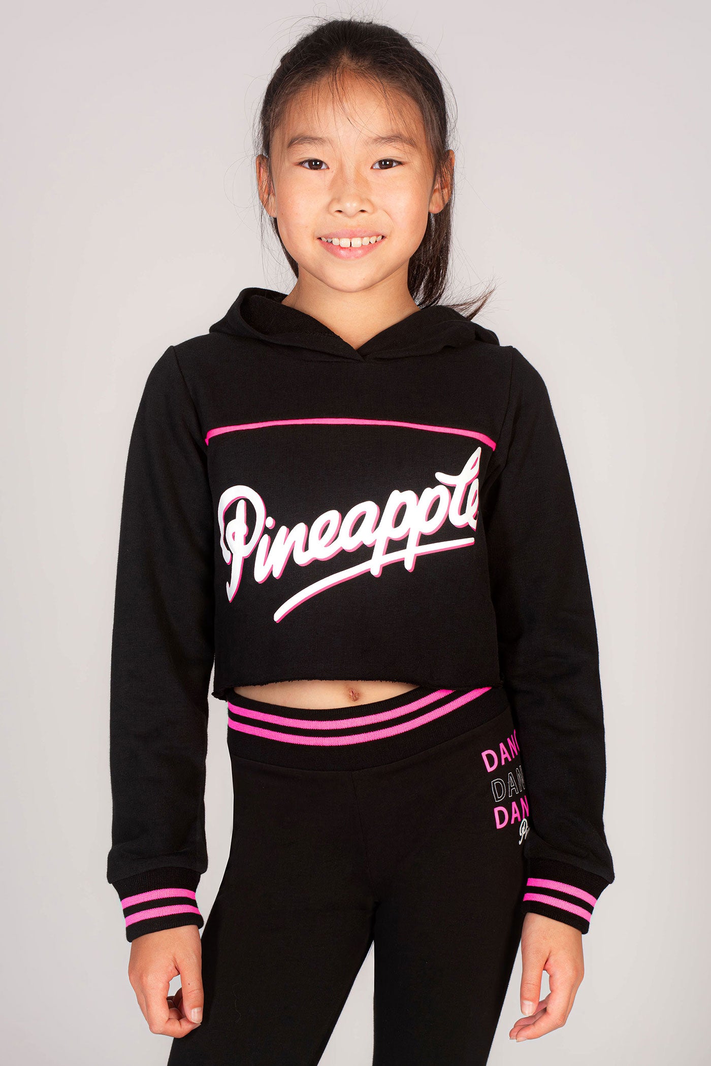 Young dancer wearing a black cropped hoodie decorated with neon pink stripes and Pineapple Dance Studios branding across the chest.