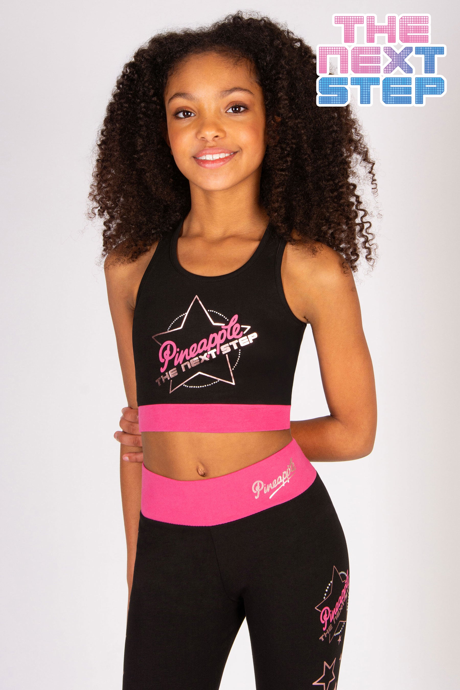 Girls' black sports crop top with The Next Step x Pineapple logo and hot pink elasticated waistband.