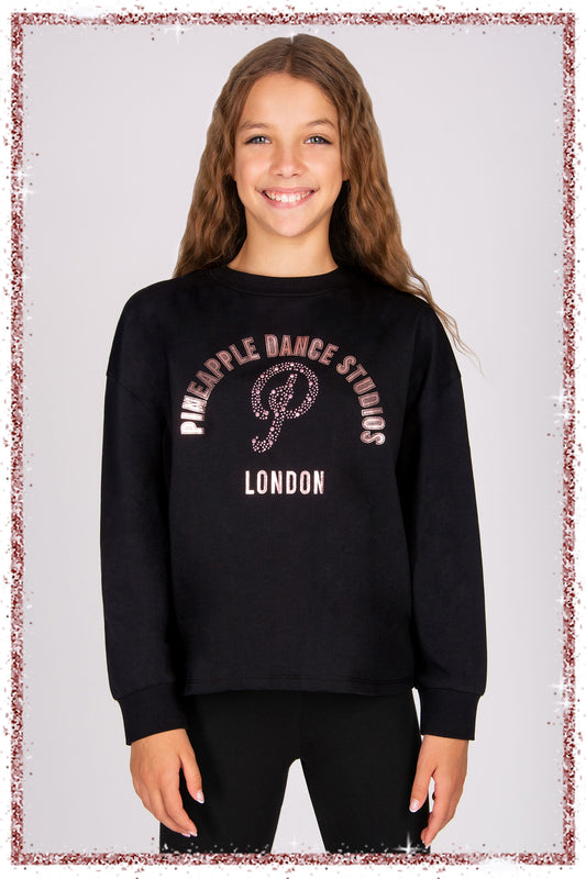 Girl wearing a black long sleeve top with rose gold foil 'Pineapple Dance Studios' print and pink diamantés on a grey background.