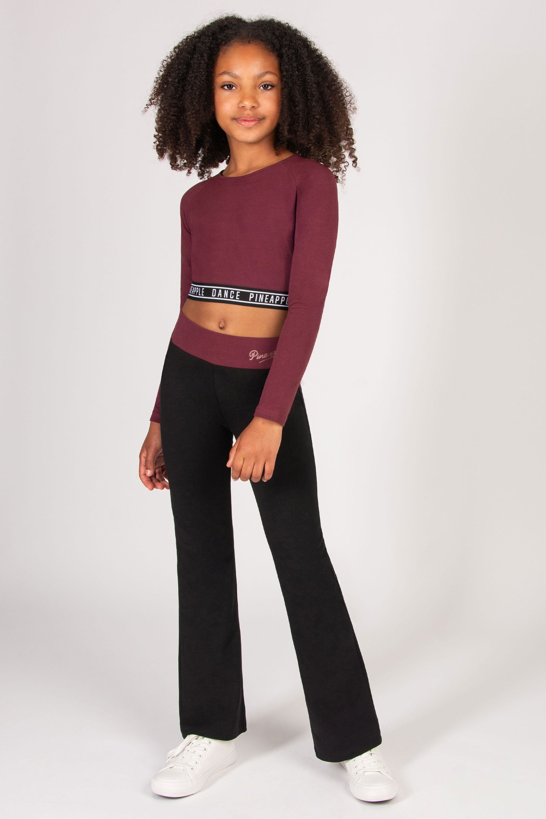 Young girl wearing a burgundy long-sleeve crop top and black flared trousers on a white background