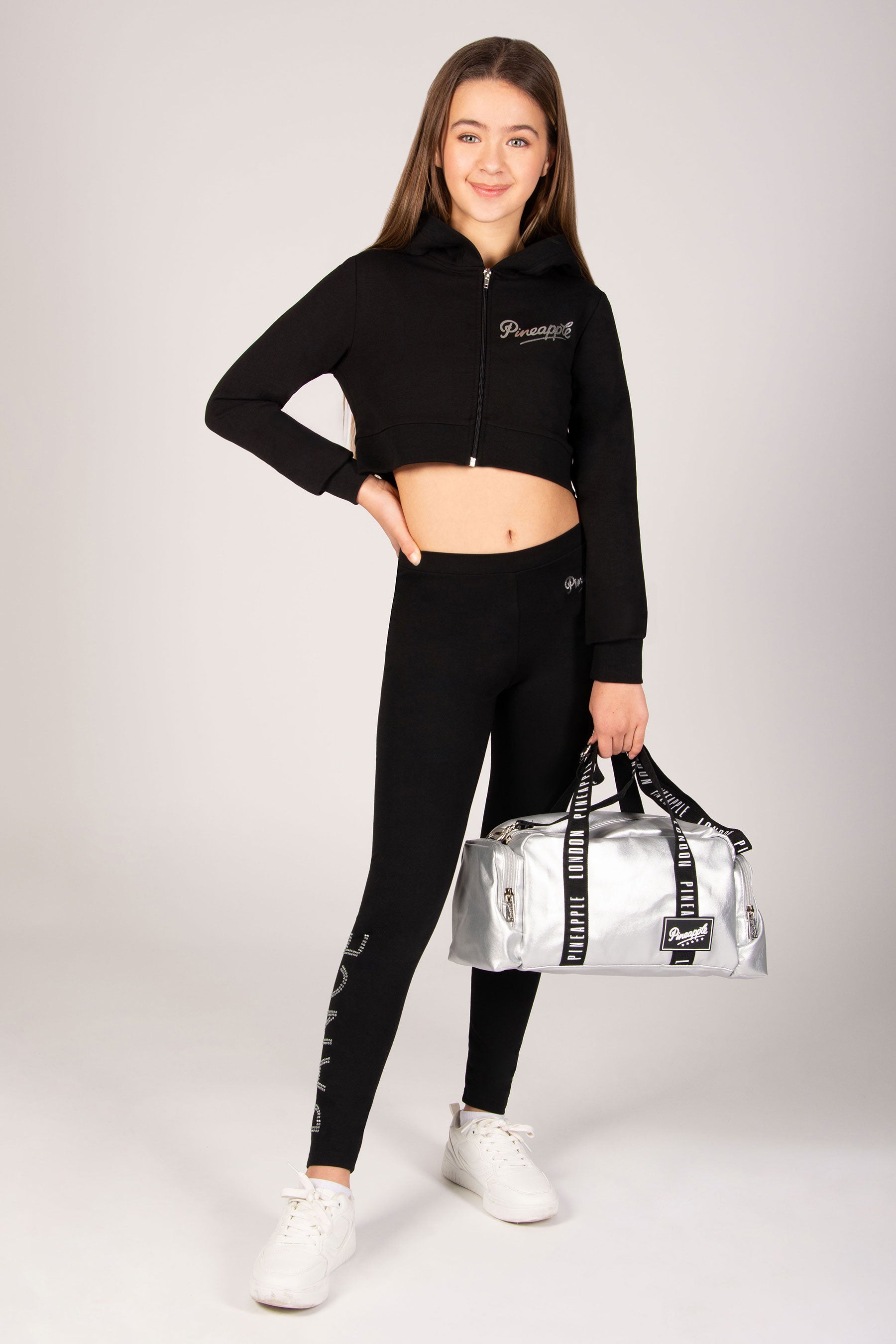 Girl in black athletic outfit holding a silver duffel bag on a white background