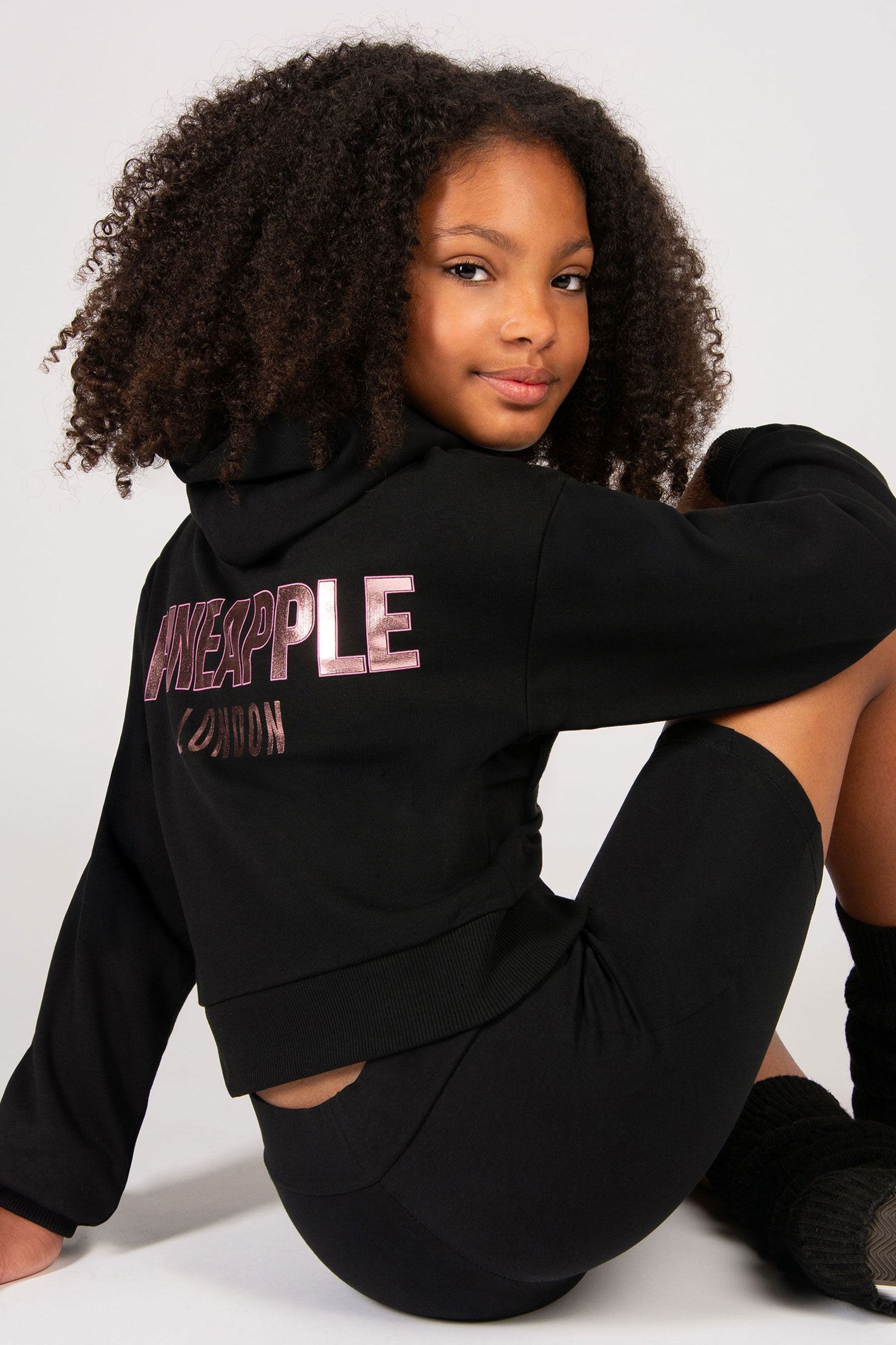 Girls wearing a black hoodie with 'PINEAPPLE' branding on the back on a white background