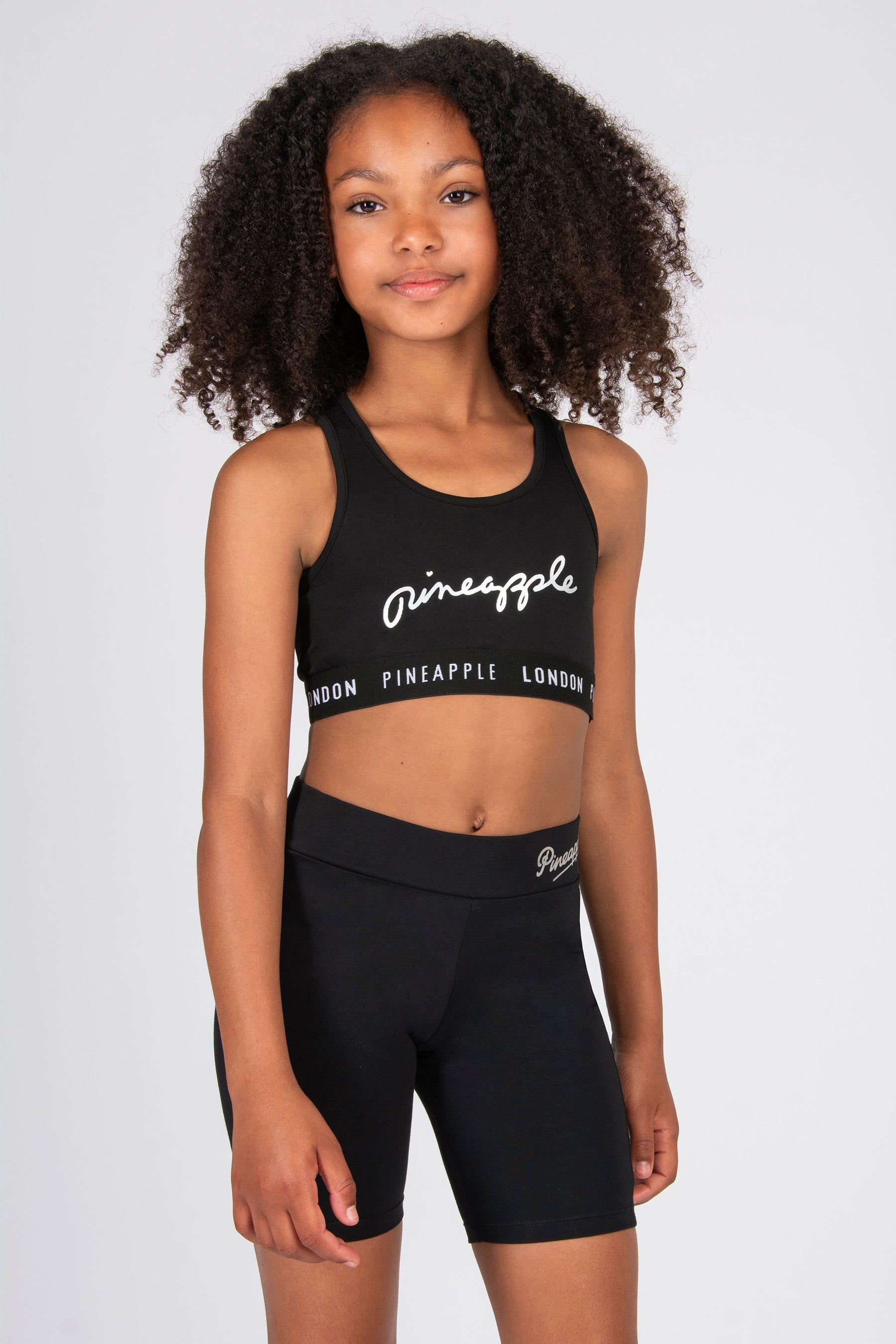 Young girl wearing a black sports bra and cycling shorts with 'Pineapple London' branding on a white background