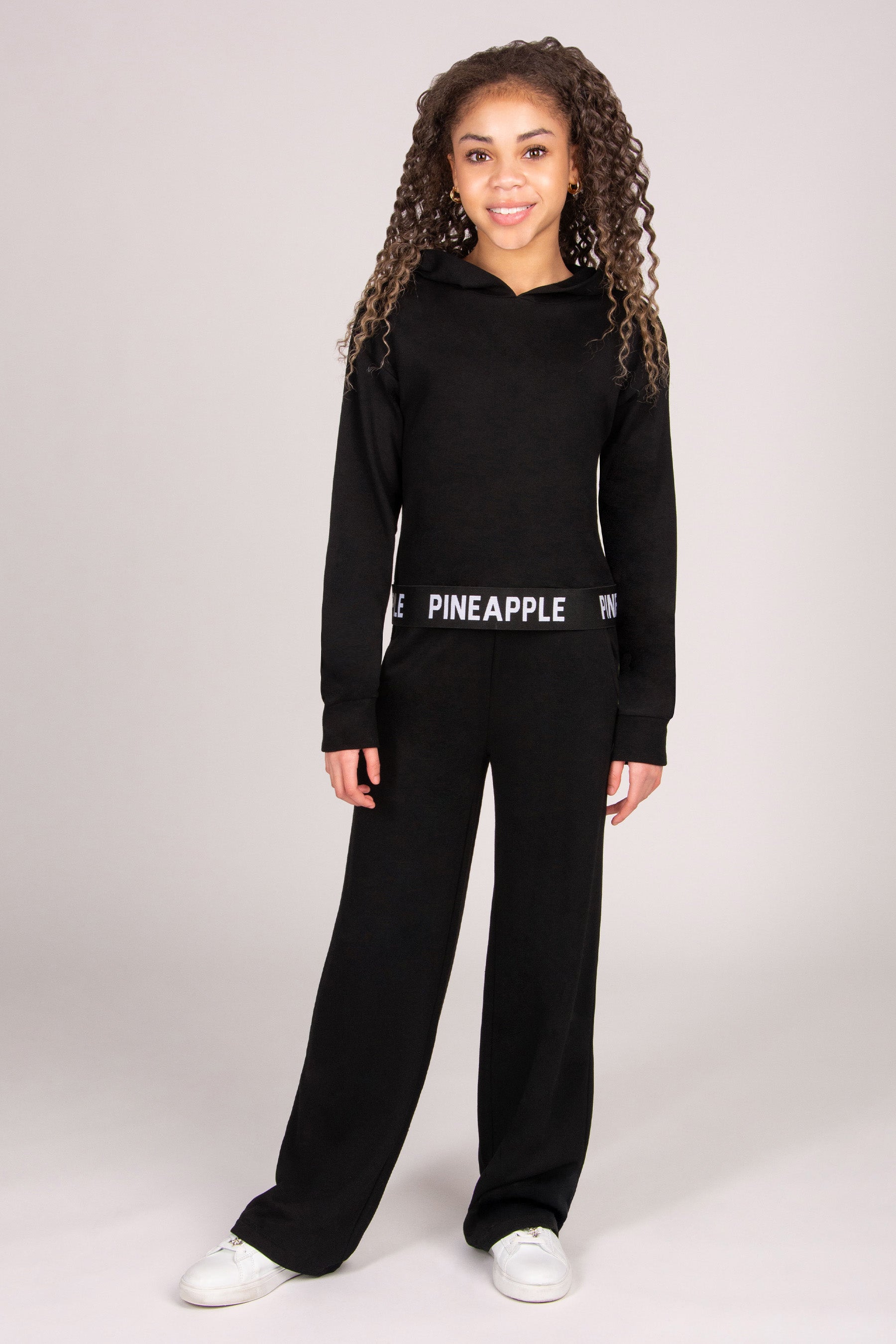 Girl wearing a black lightweight hoodie with 'Pineapple Dance' tape hem with matching black trousers.