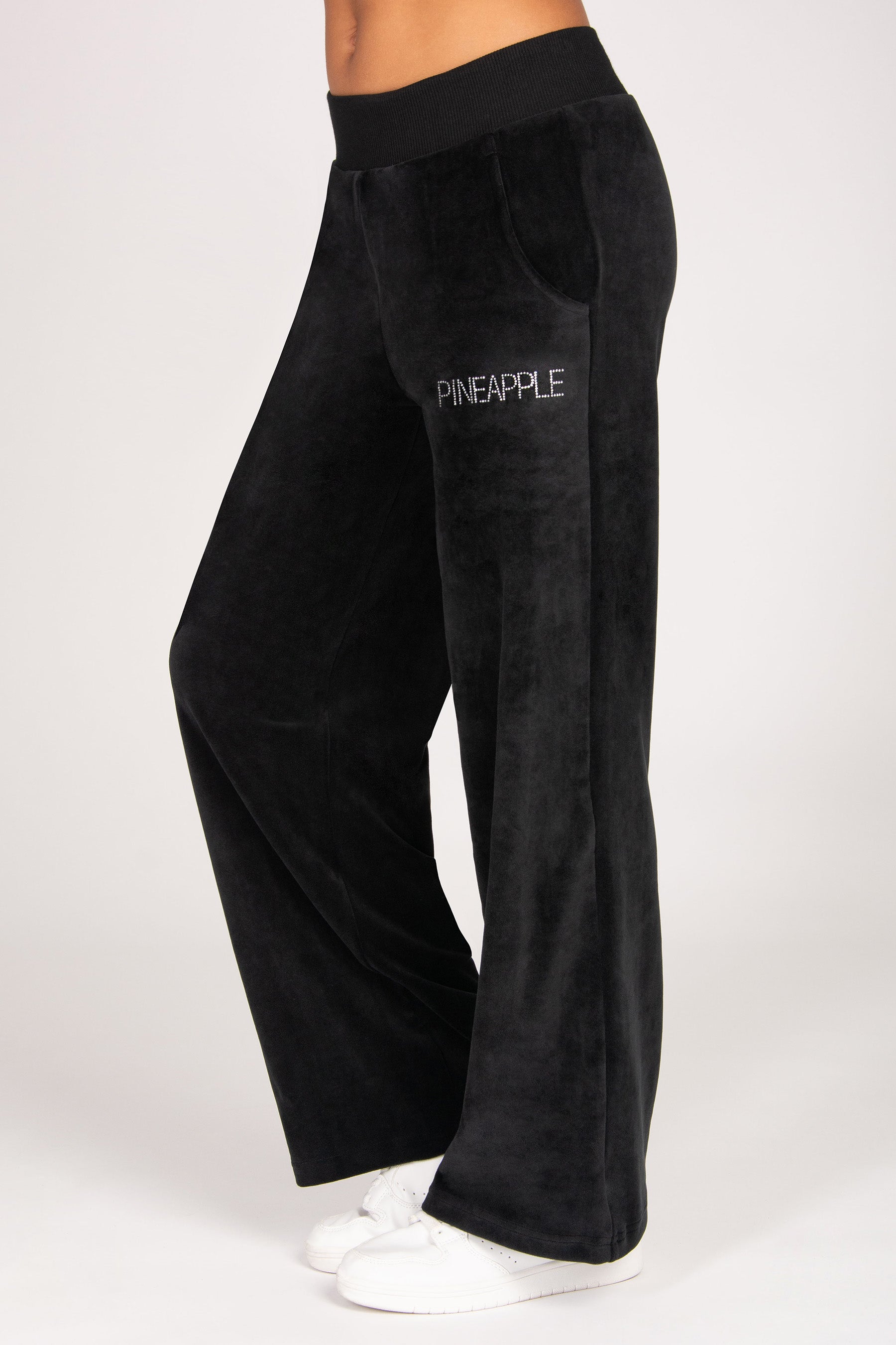 Black wide-leg velvet trousers with a small diamanté 'PINEAPPLE' logo on the left thigh, shown on a model wearing white sneakers against a light grey background.