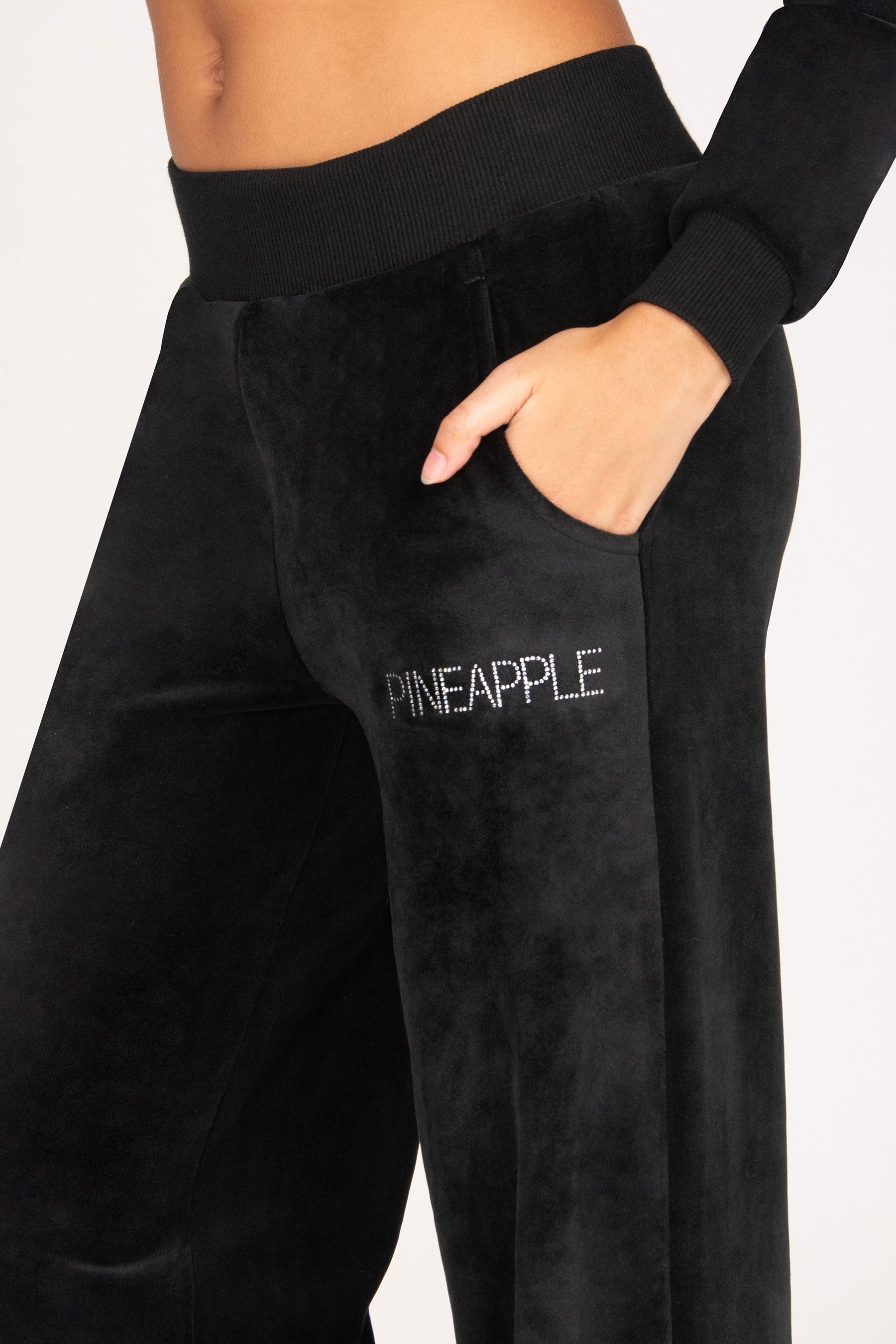 Close-up of a girl wearing black velvet trousers with a ribbed waistband and side pockets, featuring the word 'PINEAPPLE' in diamantés on the thigh.