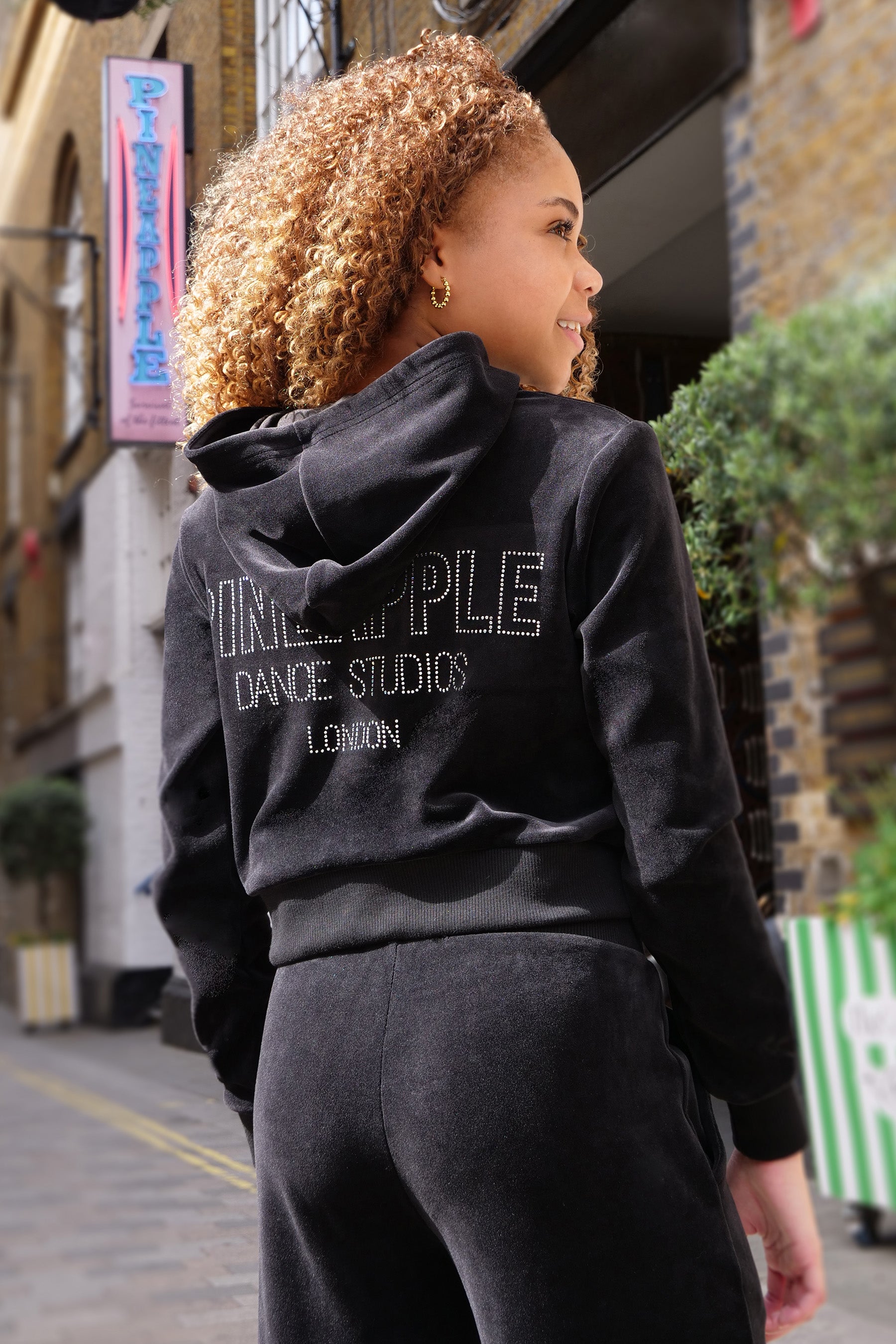 A girl shown from behind wearing a black velour zip-up hoodie and matching sweatpants. The back of the hoodie features diamanté text reading 'PINEAPPLE DANCE STUDIOS LONDON'.