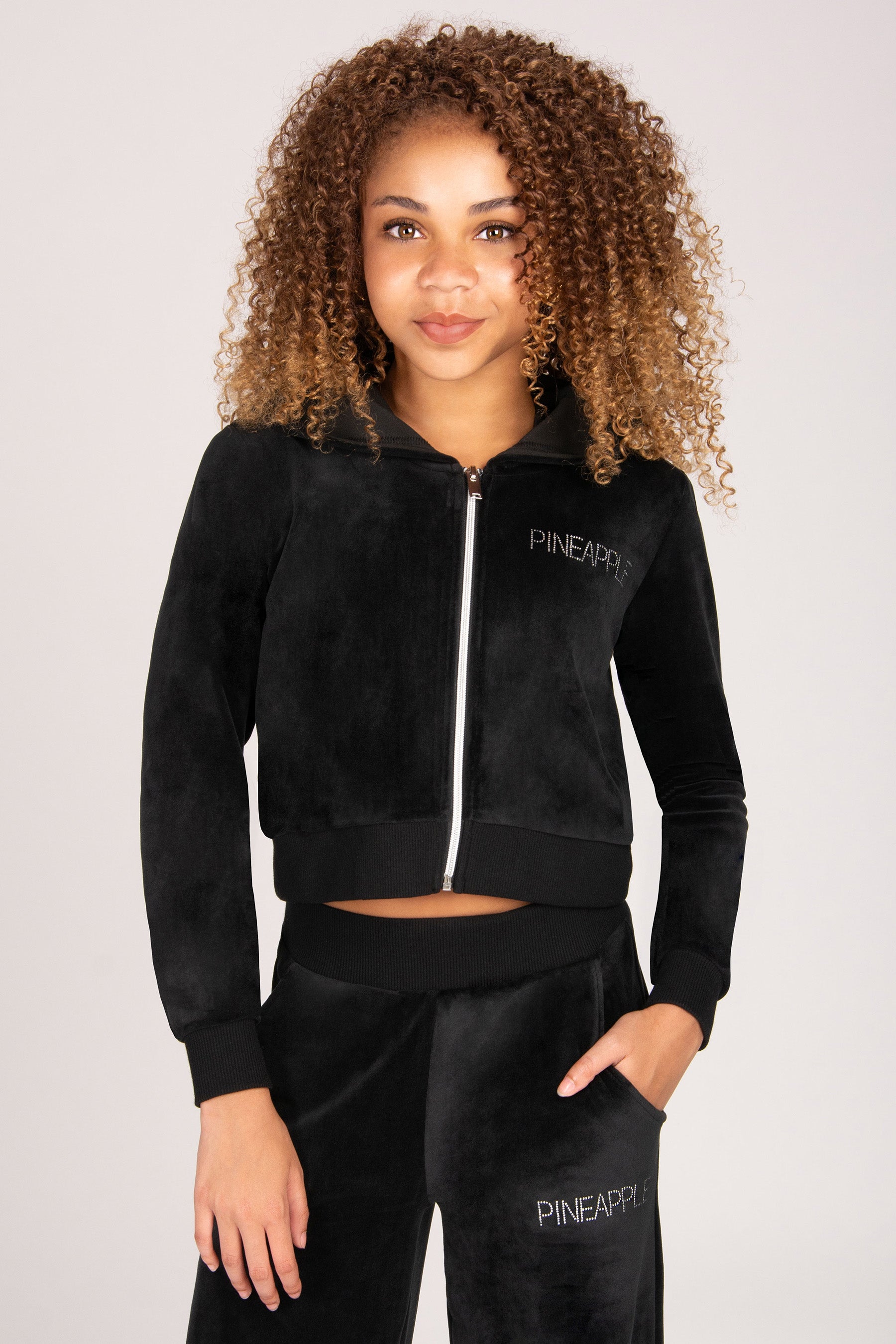 A girl wearing a black velour zip-up hoodie and matching velour sweatpants with pockets against a light grey studio background.