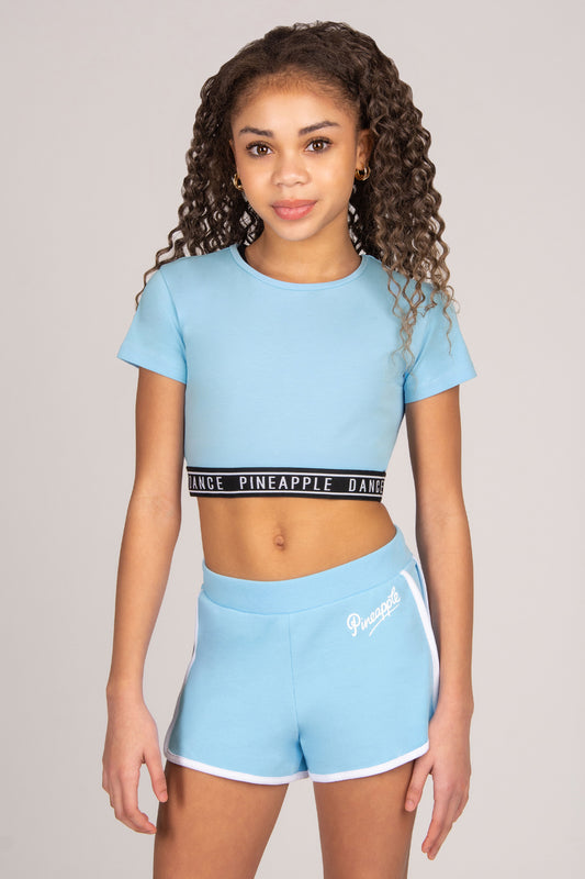 Girl wearing a light blue crop top and shorts with 'Pineapple' branding on a plain background