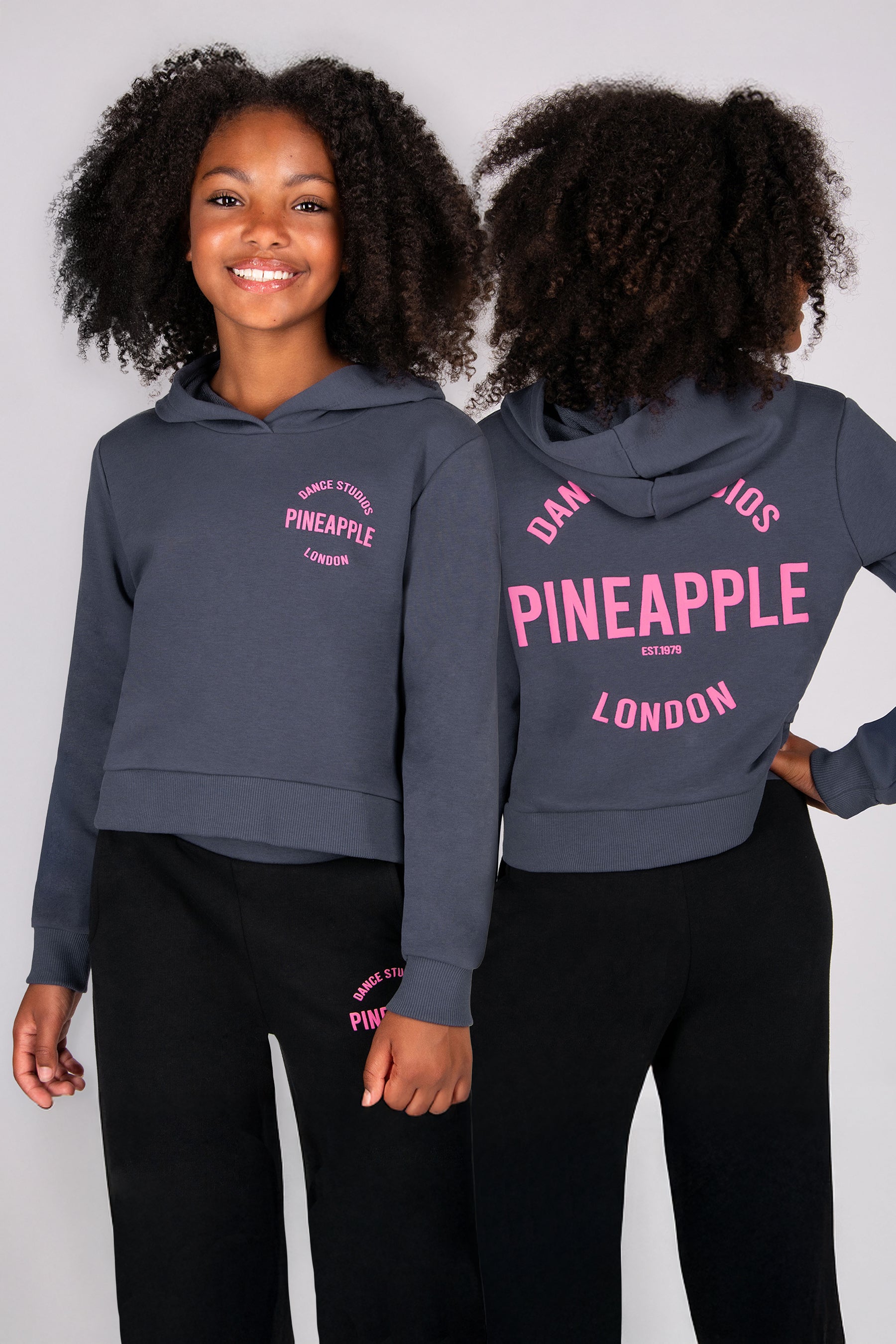 Girls wearing a dark grey hoodie with 'Pineapple London' branding on the back on a plain background