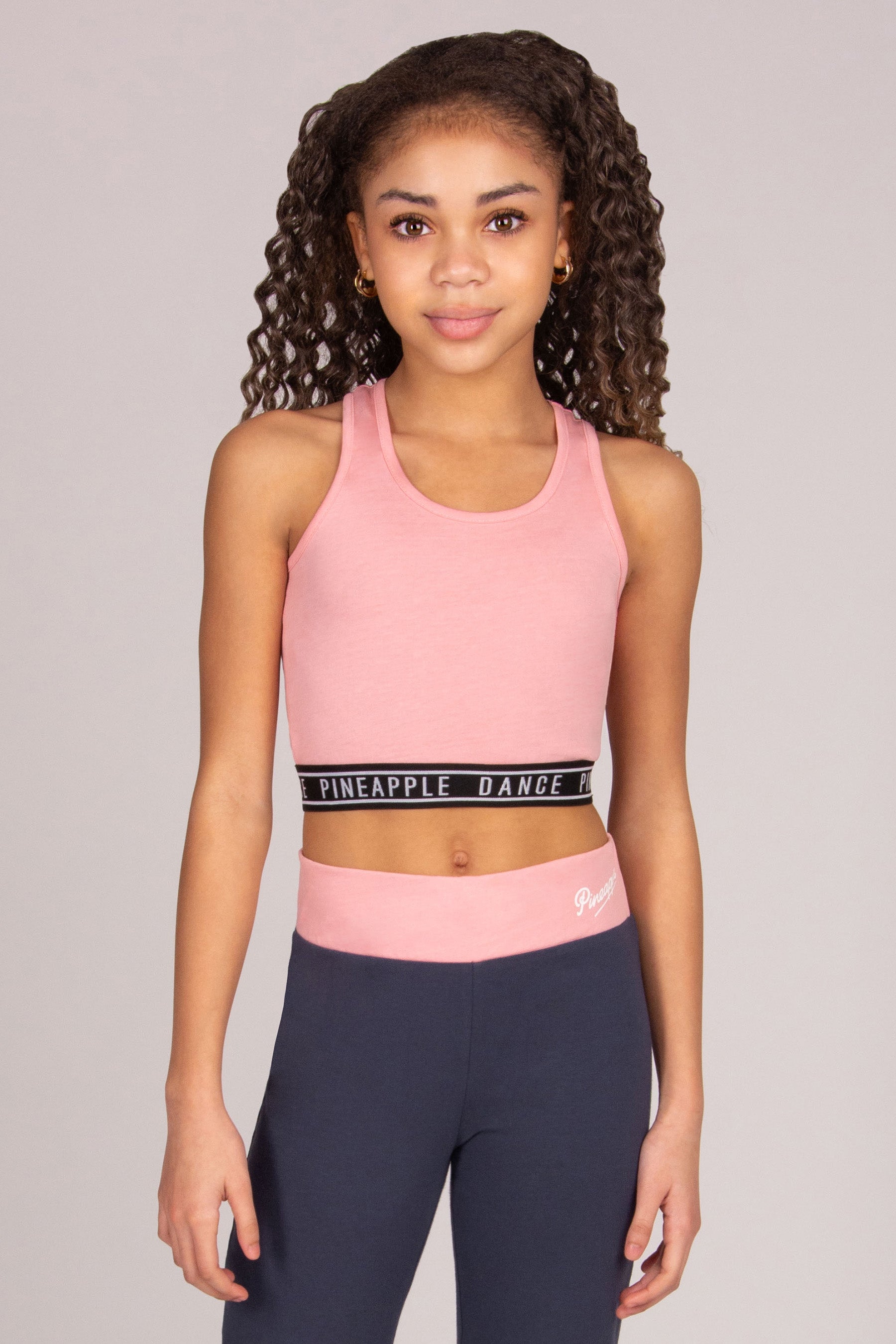 Girl wearing a pink sports crop top and dark grey leggings with 'Pineapple Dance' branding on a plain background
