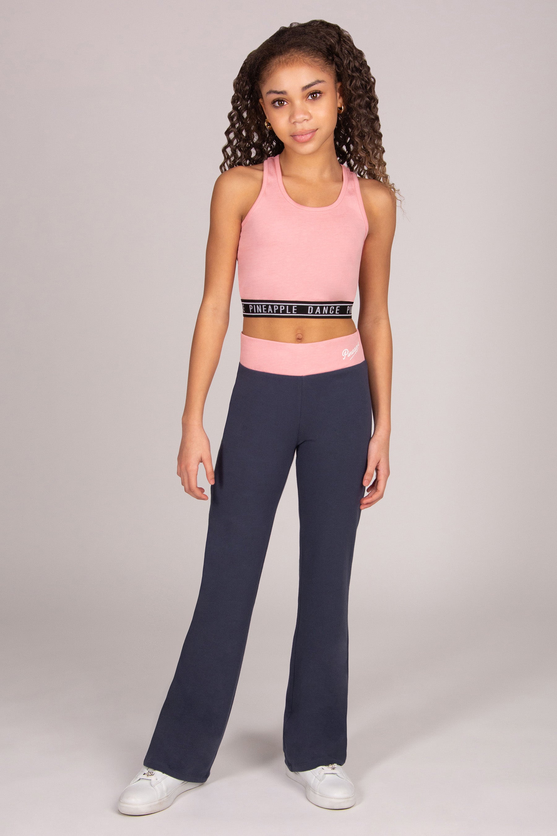 Girl wearing light pink sports crop top with matching dark grey bootcut trousers on plain background.