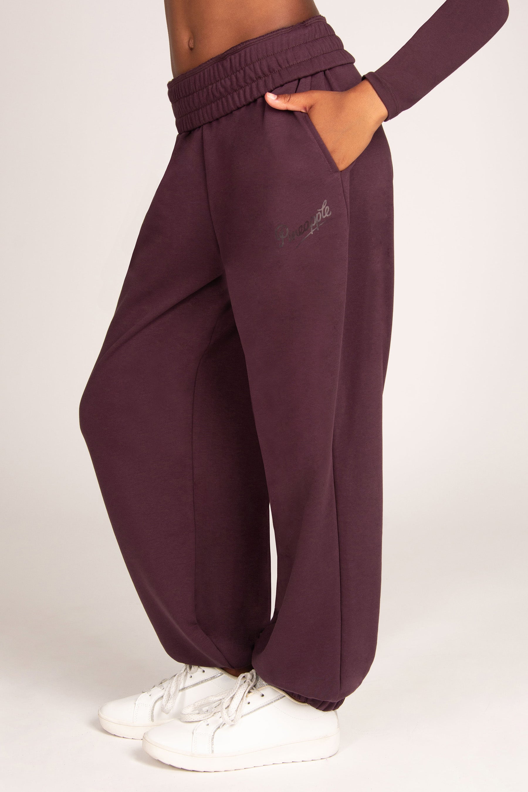 A person wearing loose, wide-leg jogger sweatpants in a deep plum purple color, paired with white sneakers, shown from the waist down against a light gray studio background.