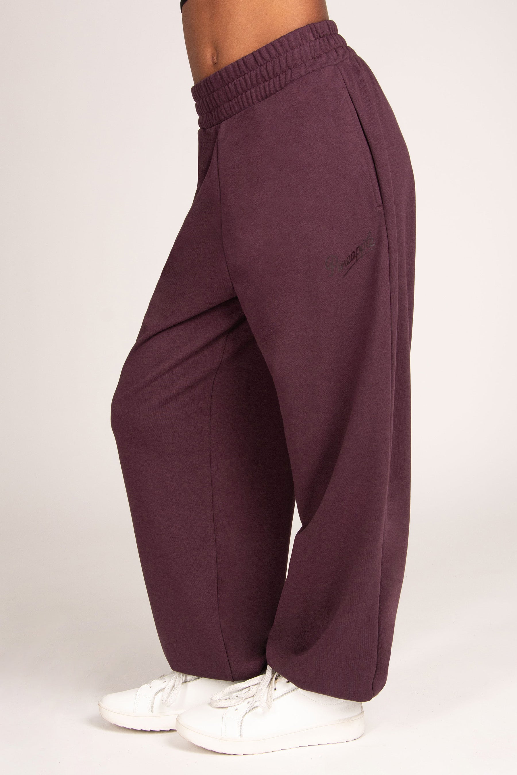 A person wearing loose, wide-leg jogger sweatpants in a deep plum purple color, paired with white sneakers, shown from a side profile against a plain light background.