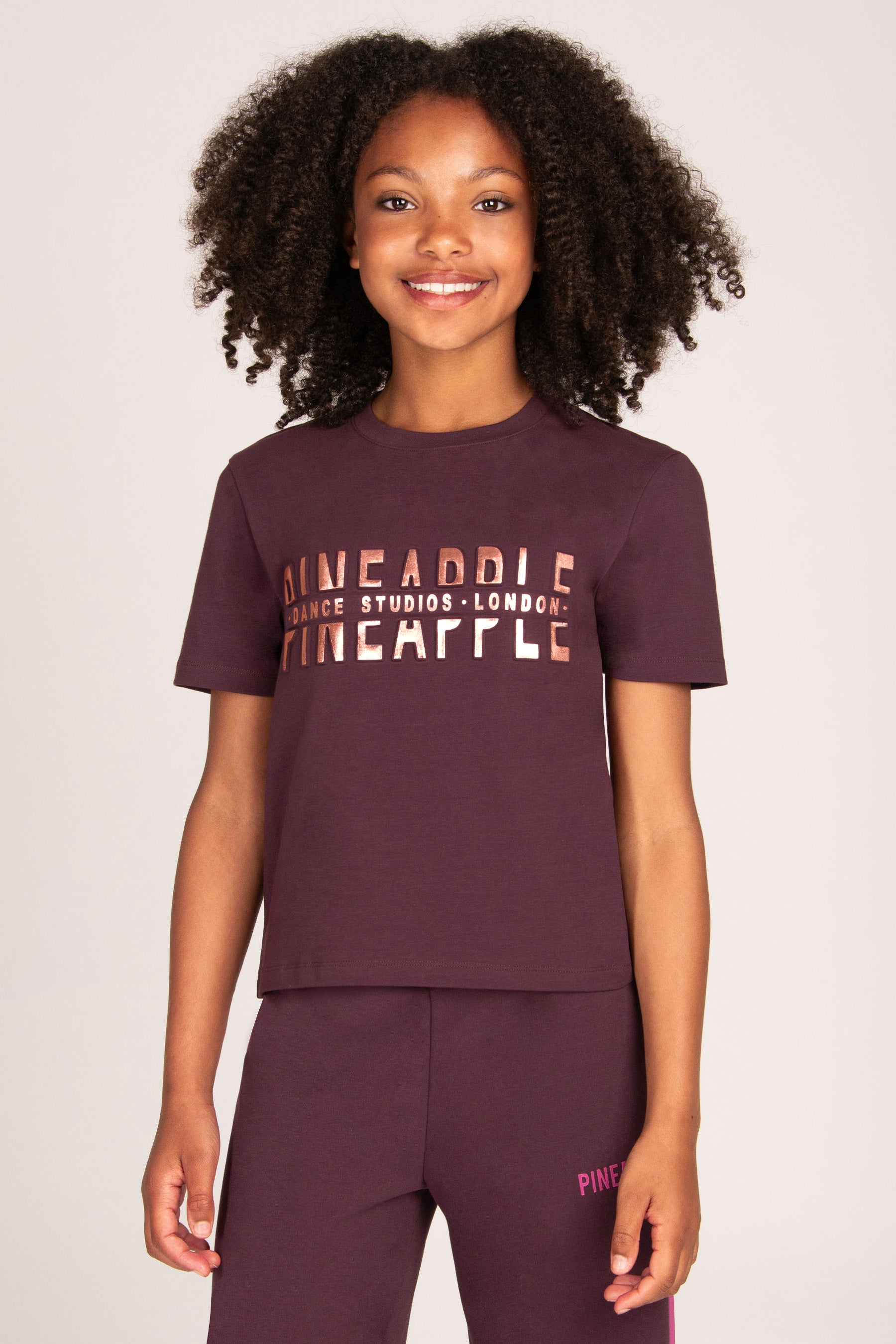 Girl wearing a maroon brown t-shirt with rose gold foil embossed 'Dance Studio London' print and matching pants on a plain background