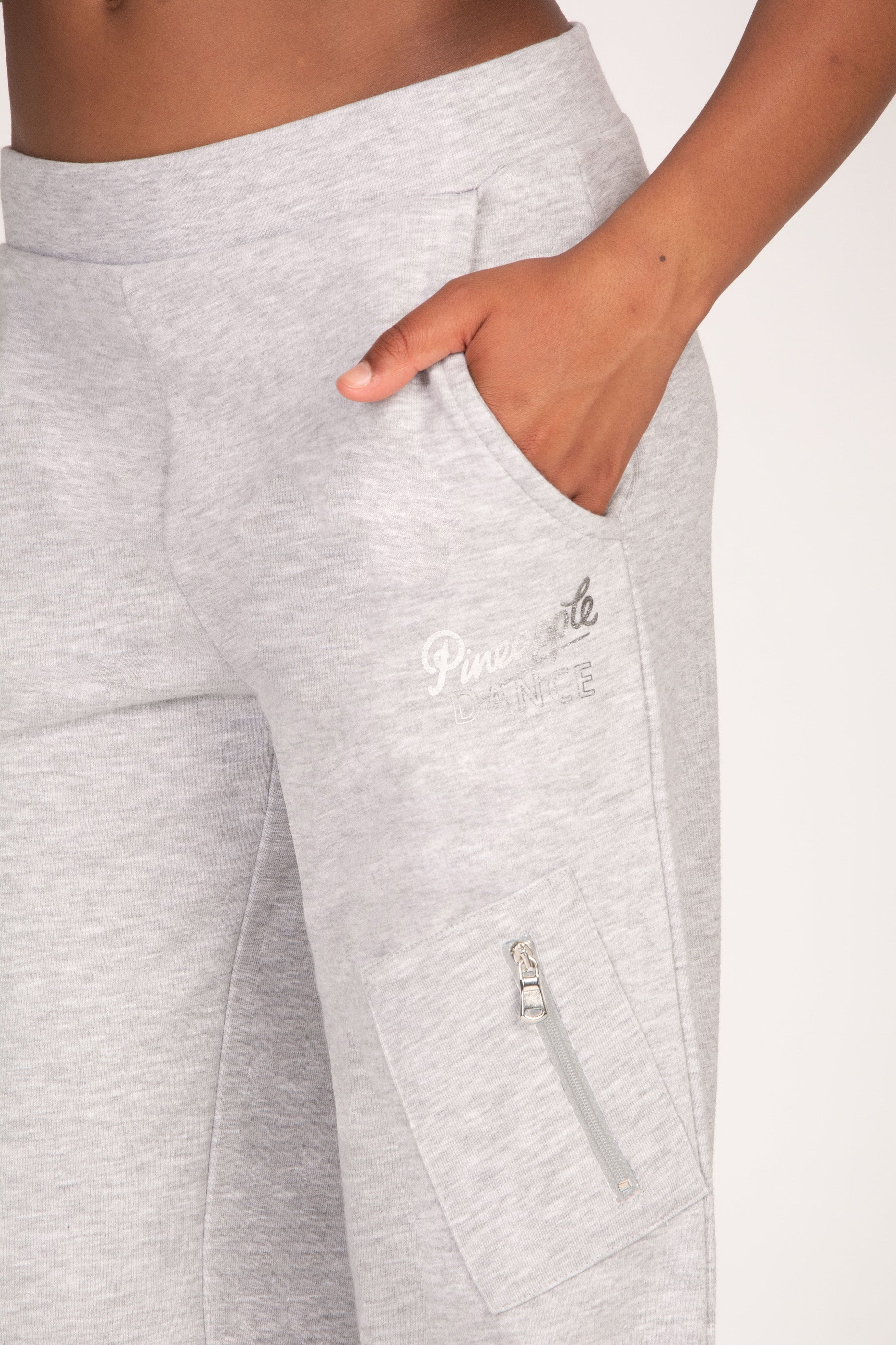 Close-up of a person wearing light grey jogger sweatpants, showing a side pocket with a hand in the pocket and a small silver foil logo on the thigh.