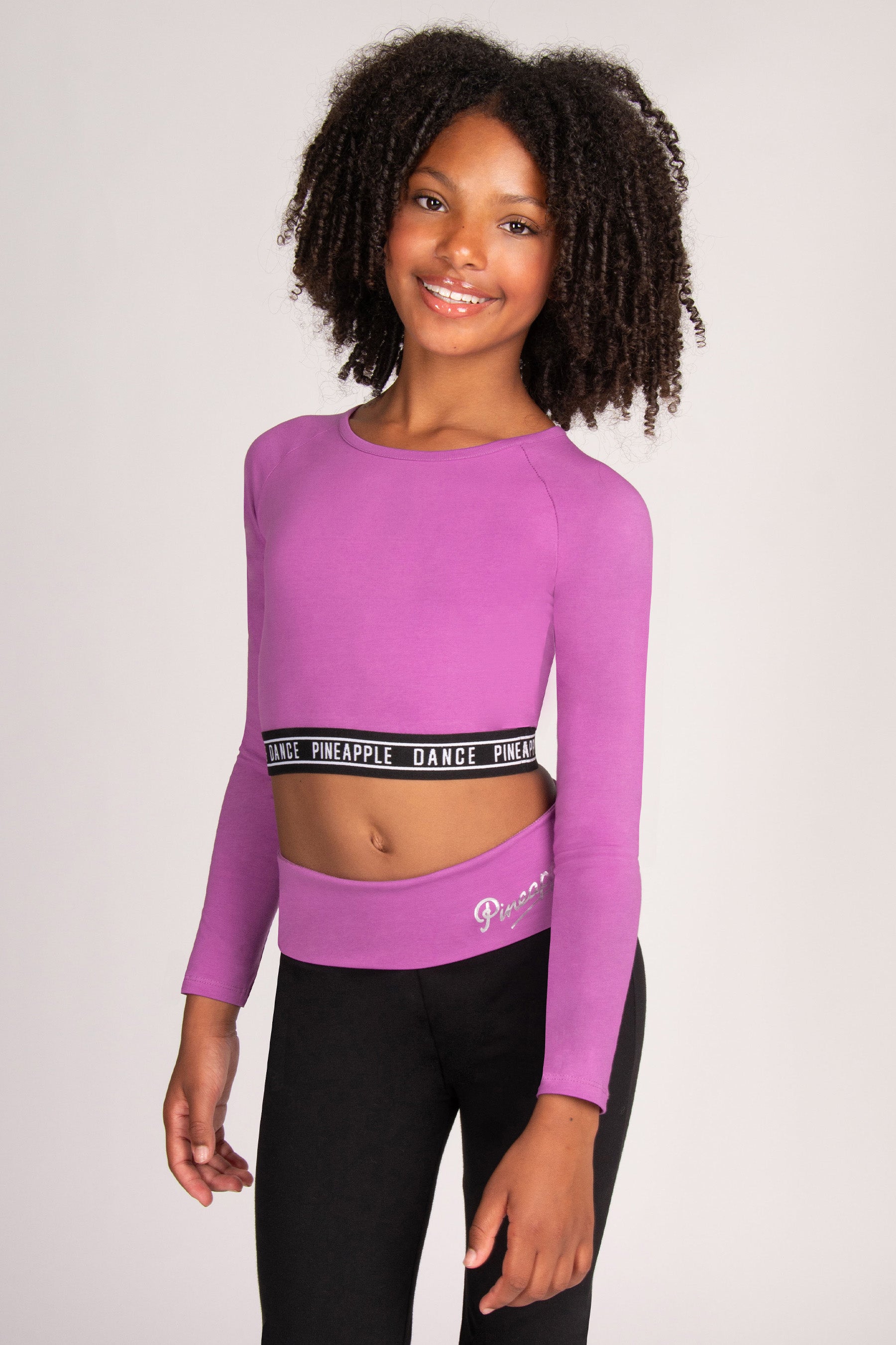 A young girl with curly black hair wearing a bright purple long-sleeve crop top with a black branded waistband and black leggings, standing against a light grey studio background.