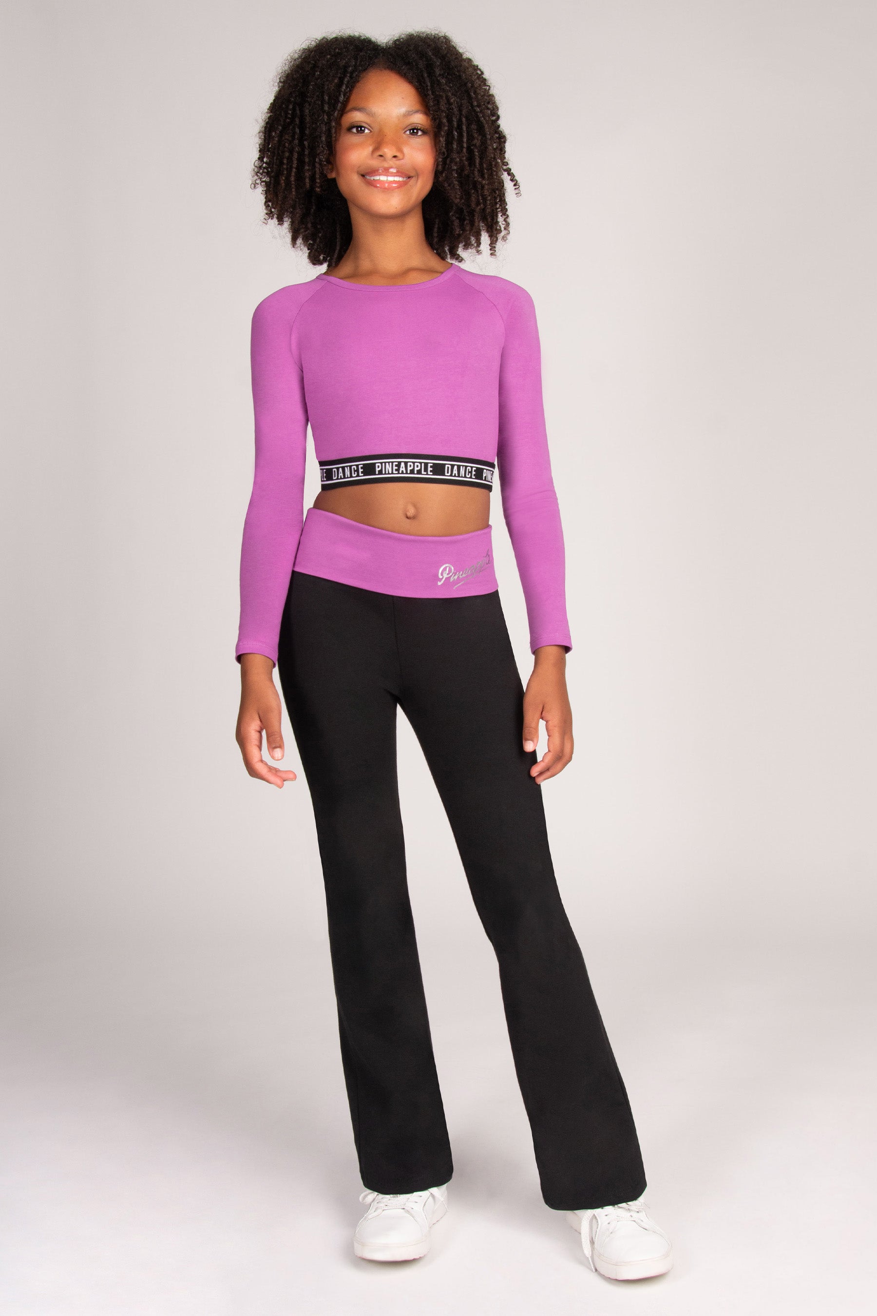 A young girl modeling a two-piece activewear set consisting of a long-sleeve crop top in bright purple with a black and white waistband, and black wide-leg pants, paired with white sneakers, against a light grey studio background.