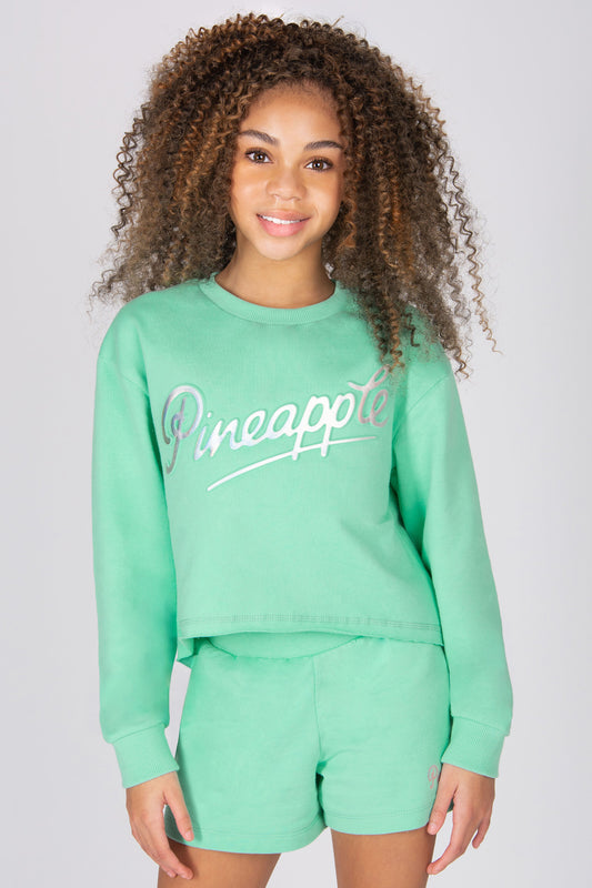 Girl wearing a mint green 'Pineapple' logo sweatshirt and sporty shorts set on a light grey background