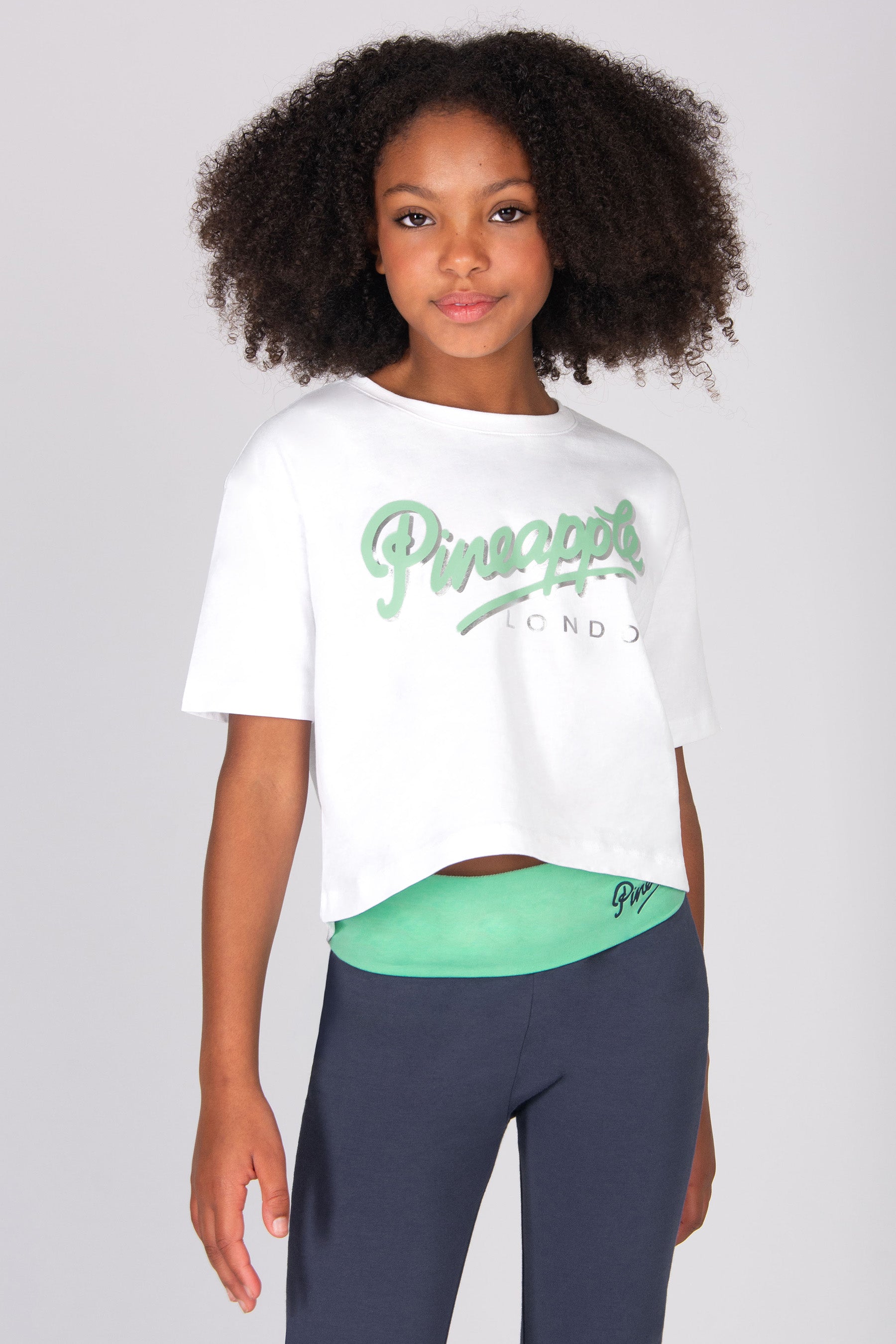 Young girl wearing a white t-shirt with light blue 'Pineapple' logo and dark grey yoga pants on a light grey background