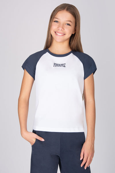 Teenage girl wearing a white and dark grey raglan sleeve t-shirt with 'Pineapple' logo on a plain background