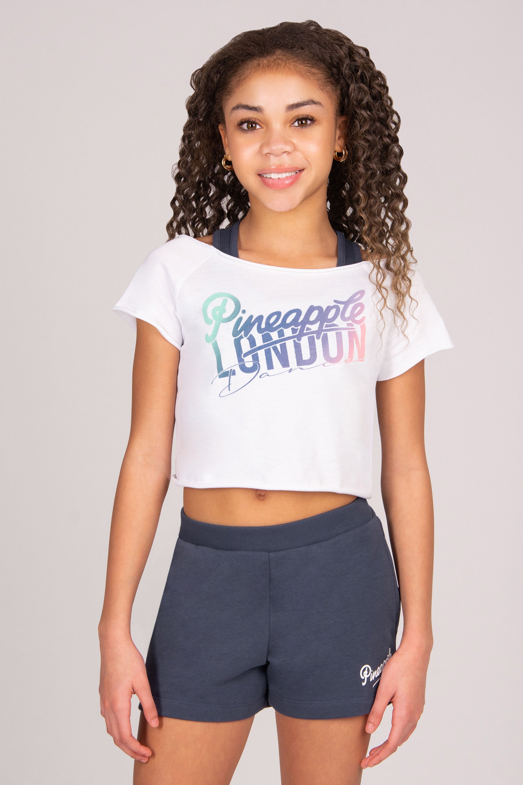 Girl wearing a white crop top with "Pineapple London Dance" print and dark grey shorts on a plain background