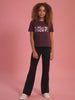 Girl wearing a maroon brown t-shirt with rose-gold embossed print and black fared trousers.