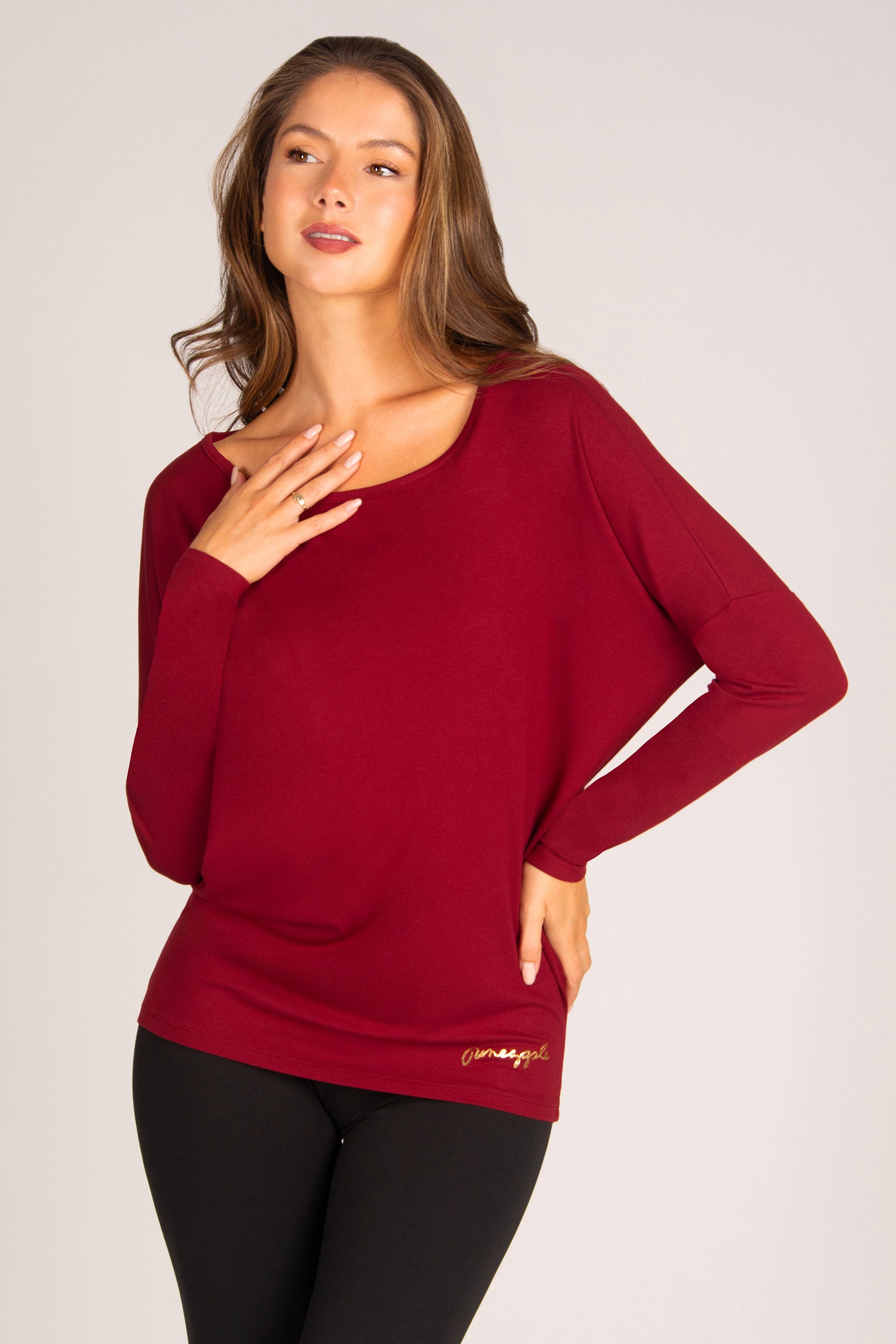 A woman modeling a long-sleeve boat-neck sweater in deep burgundy red, paired with black leggings, against a light grey studio background.