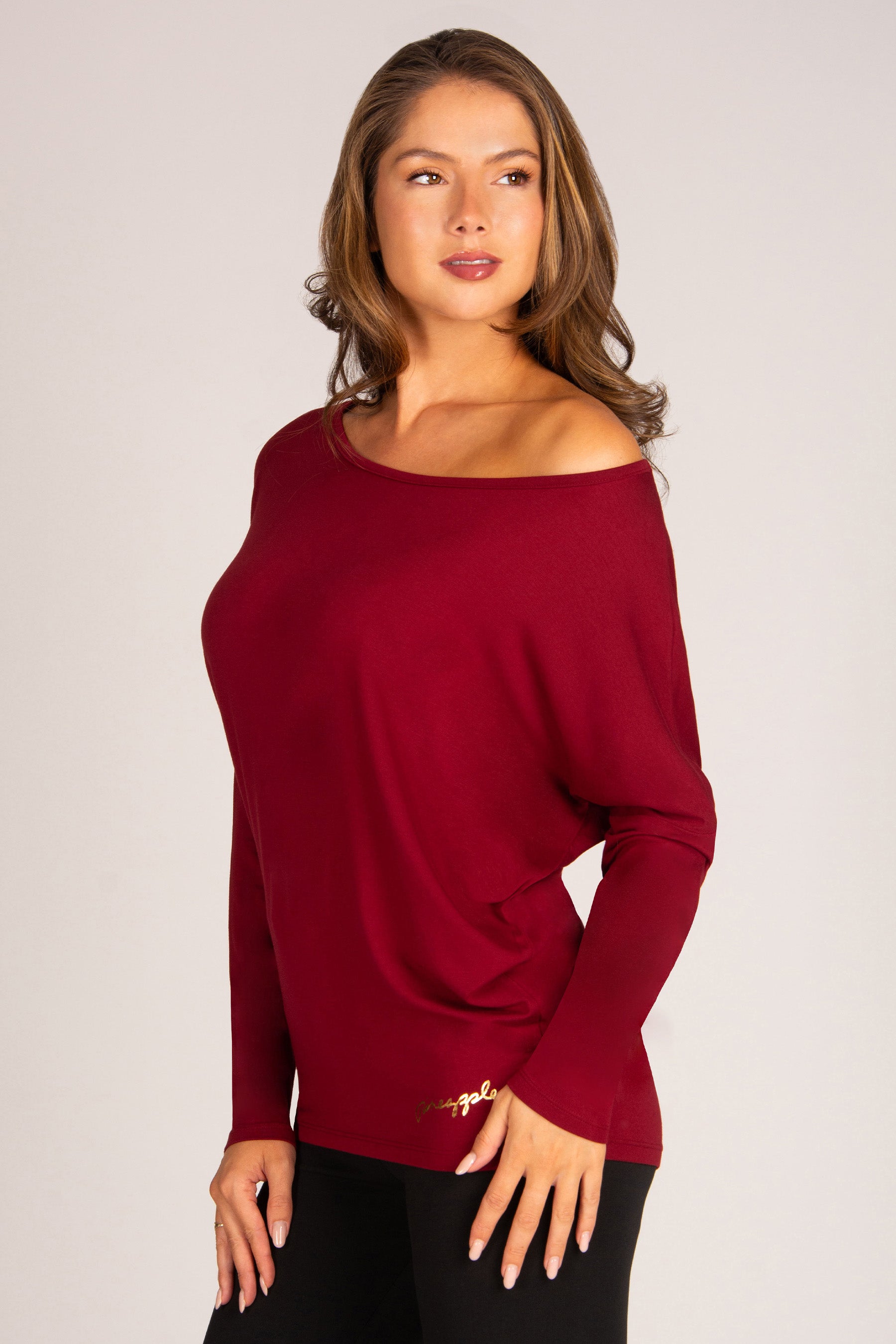 A woman modeling a long-sleeve off-the-shoulder knit top in deep burgundy, paired with black pants, against a solid light gray studio background.
