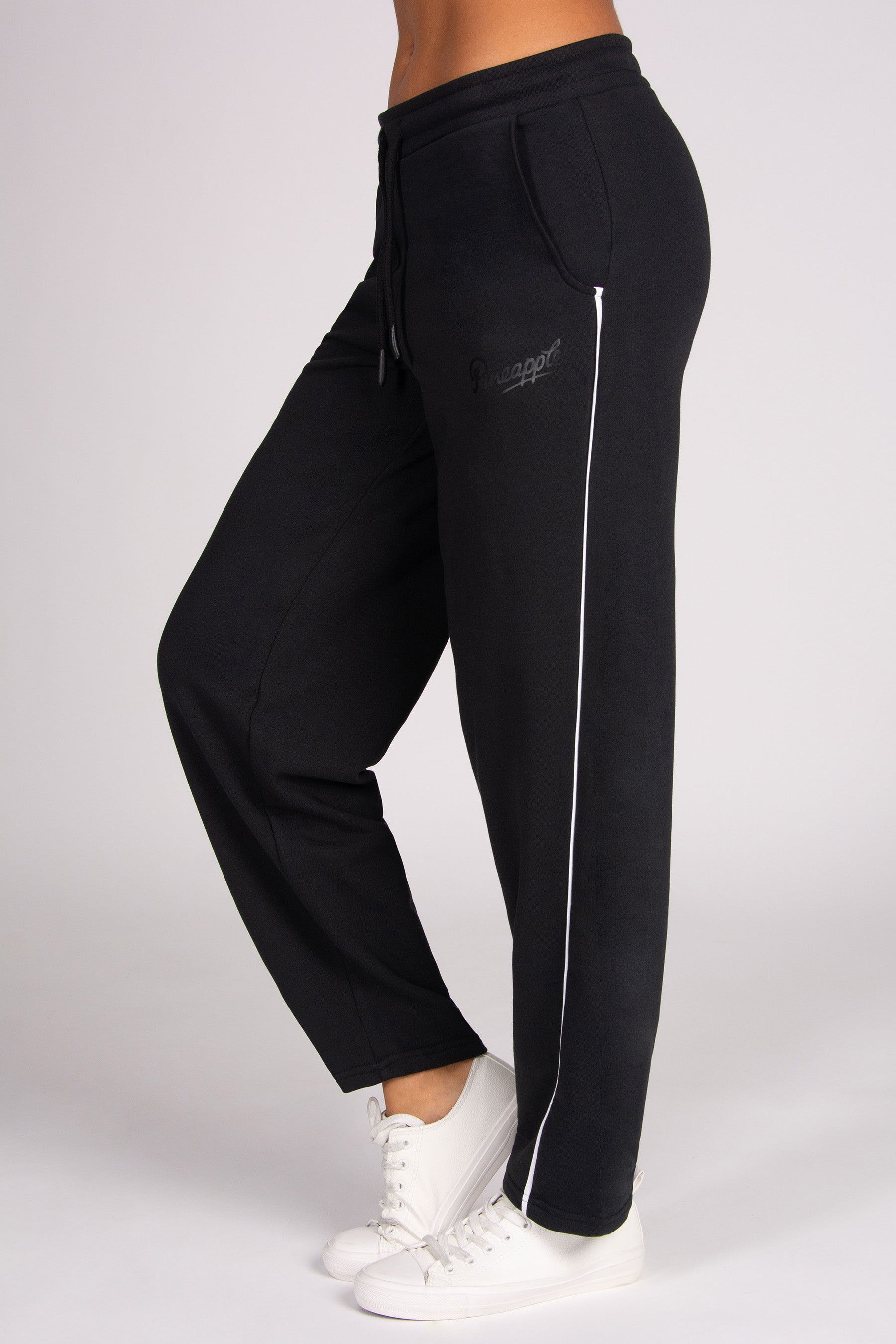 Black barrel sweatpants with a white side stripe and a small black logo on the upper thigh, shown on a model wearing white sneakers against a light grey background.