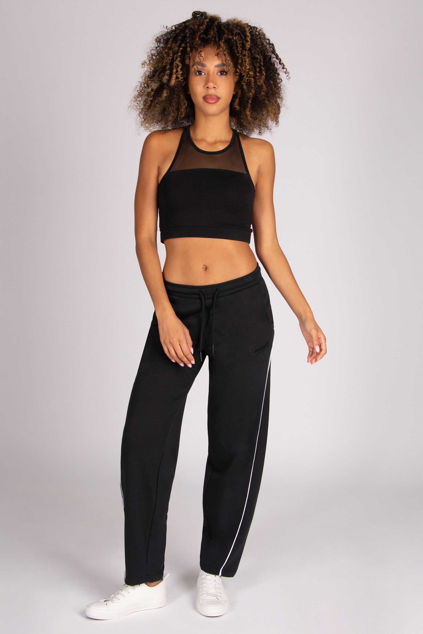A woman modelling a black athletic set consisting of a mesh-shoulder crop top and matching drawstring barrel sweatpants with a white side stripe against a light grey studio background.