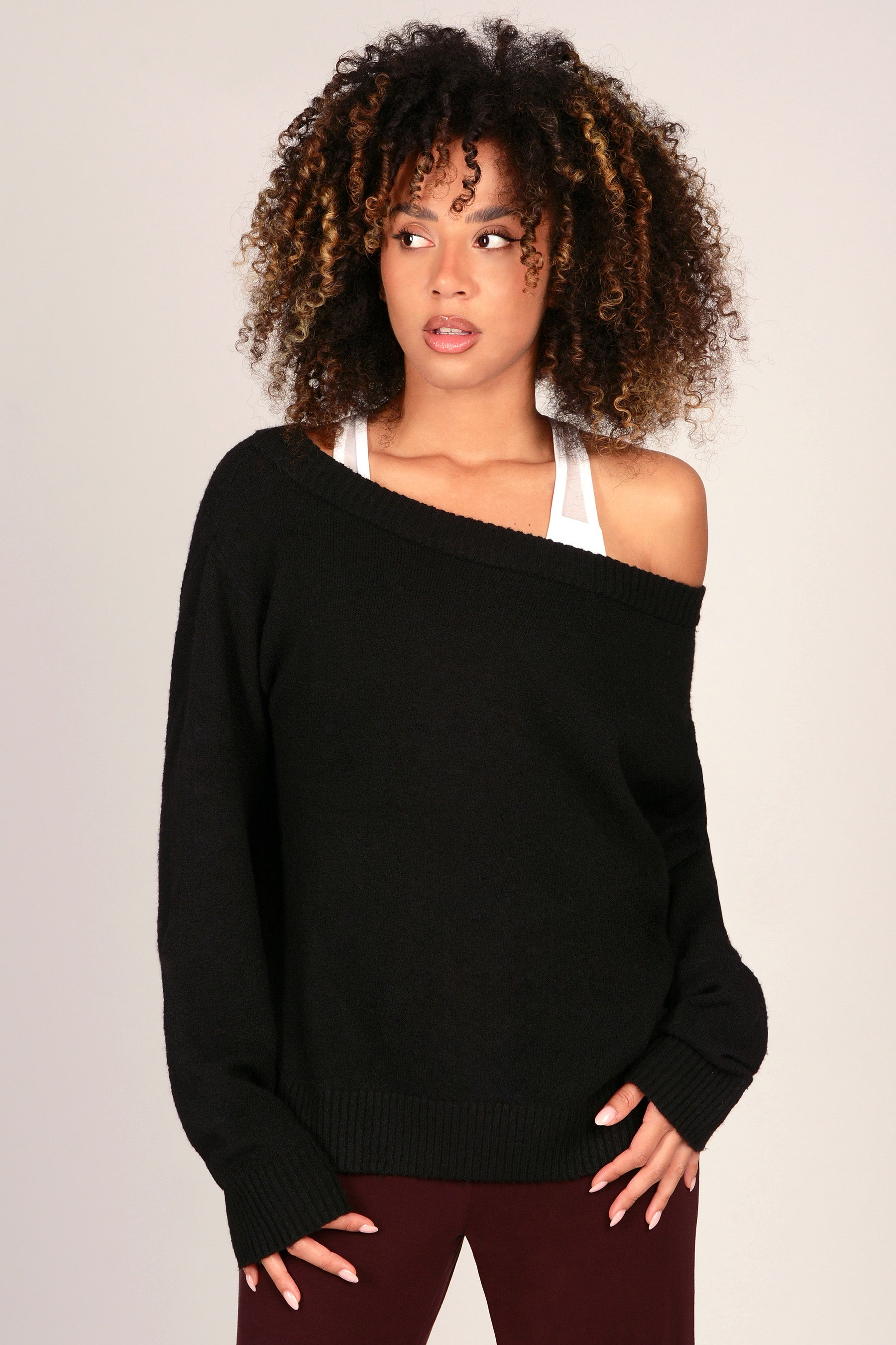 A woman wearing a black off-the-shoulder knit sweater over a white tank top and burgundy trousers, photographed against a plain light grey studio background.