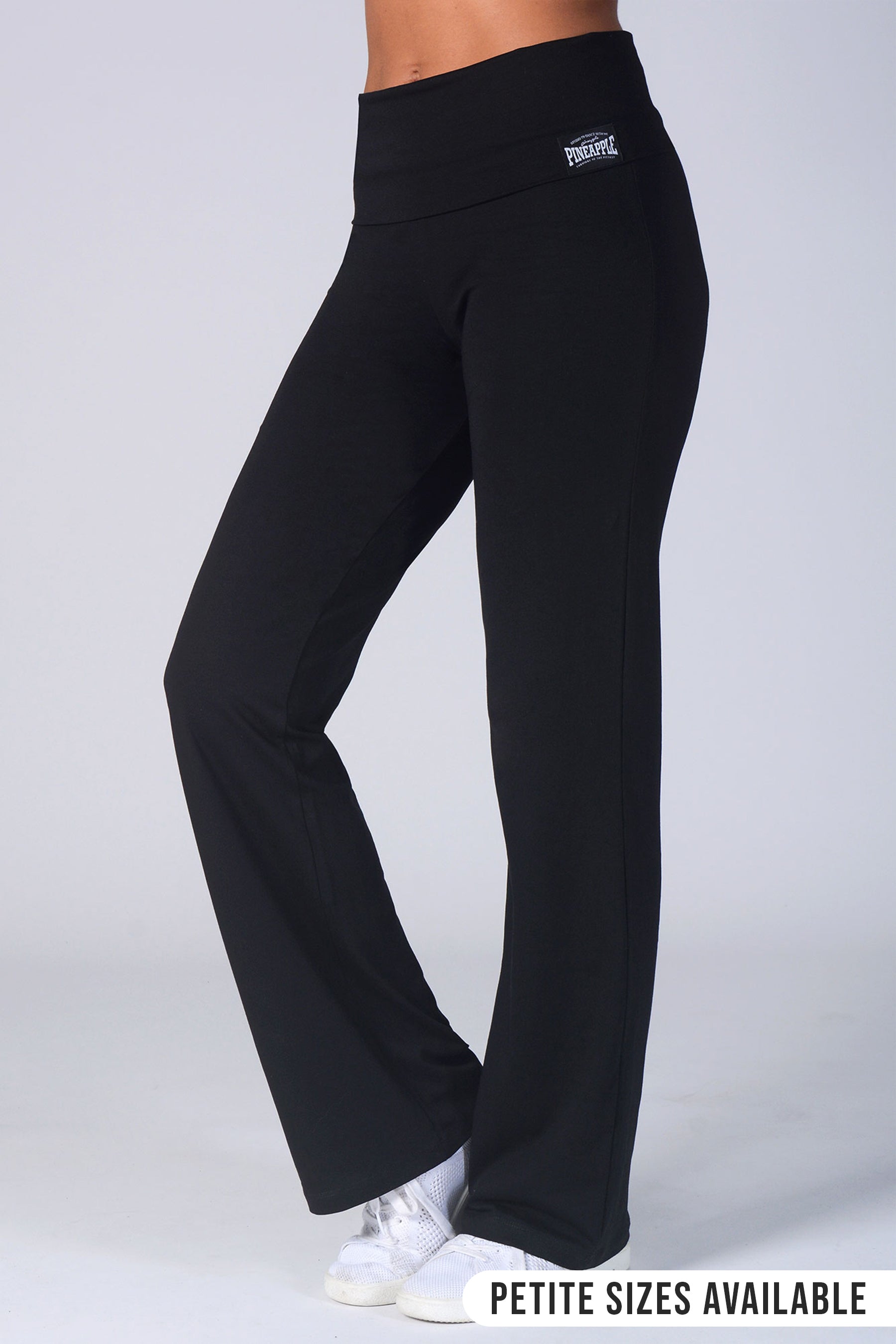 Black high-waisted yoga pants with a small white logo on the left hip, shown on a model from the waist down wearing white sneakers against a light gray background.