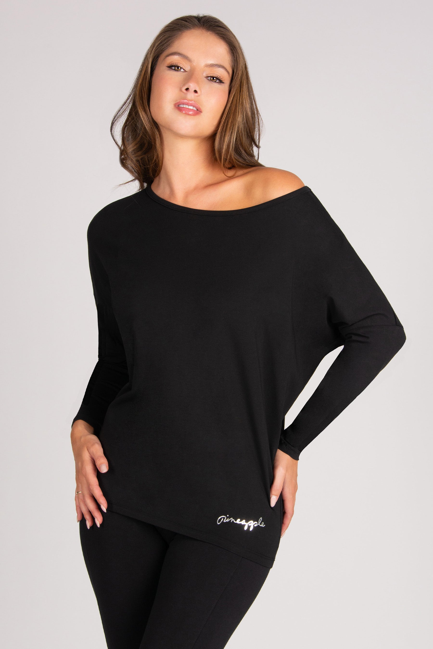 A woman modelling a black off-the-shoulder long-sleeve top with a small silver script logo near the hem, paired with black leggings, against a light grey studio background.