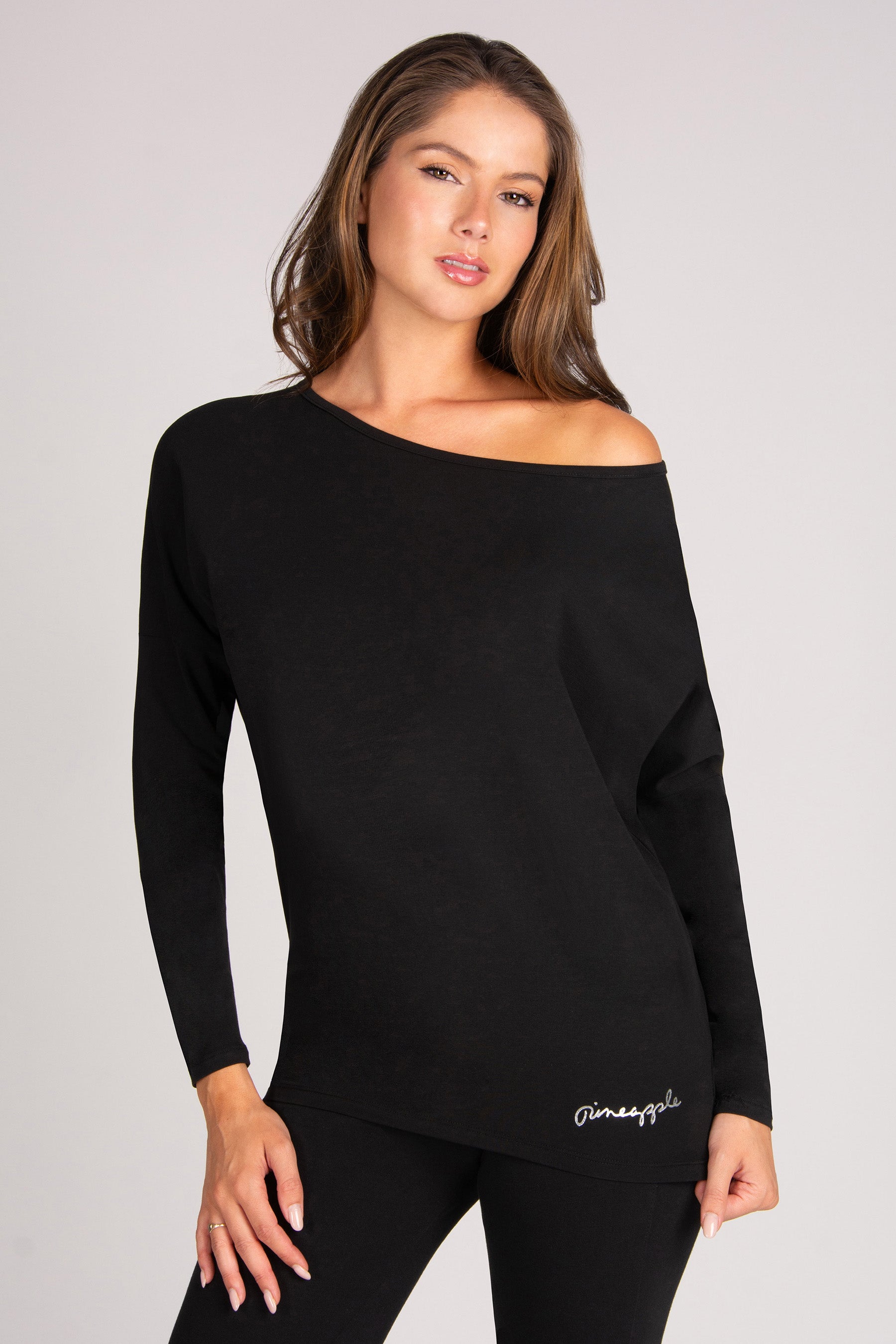 A woman modelling a black off-the-shoulder long-sleeve top with a small silver foil script logo near the lower hem, paired with black leggings, against a light grey studio background.