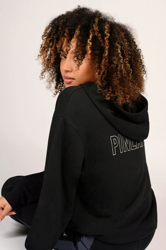 Woman wearing a black hoodie with 'PINEAPPLE' branding on the back, sitting against a white background.
