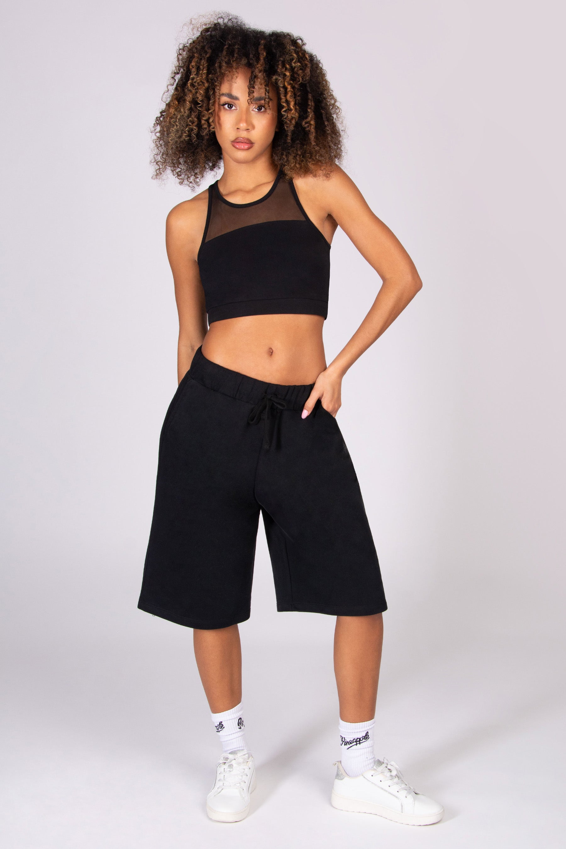 Woman wearing a black crop top and black super oversized basketball shorts  on a light grey background
