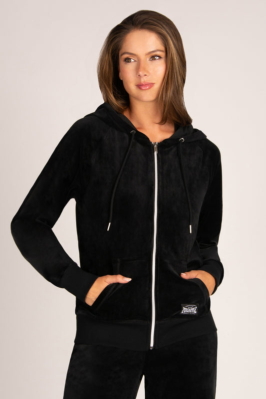 A woman modelling a black zip-up hoodie with a plush velvet-like texture, featuring a silver zipper, black drawstrings, and front pockets, set against a plain light grey studio background.