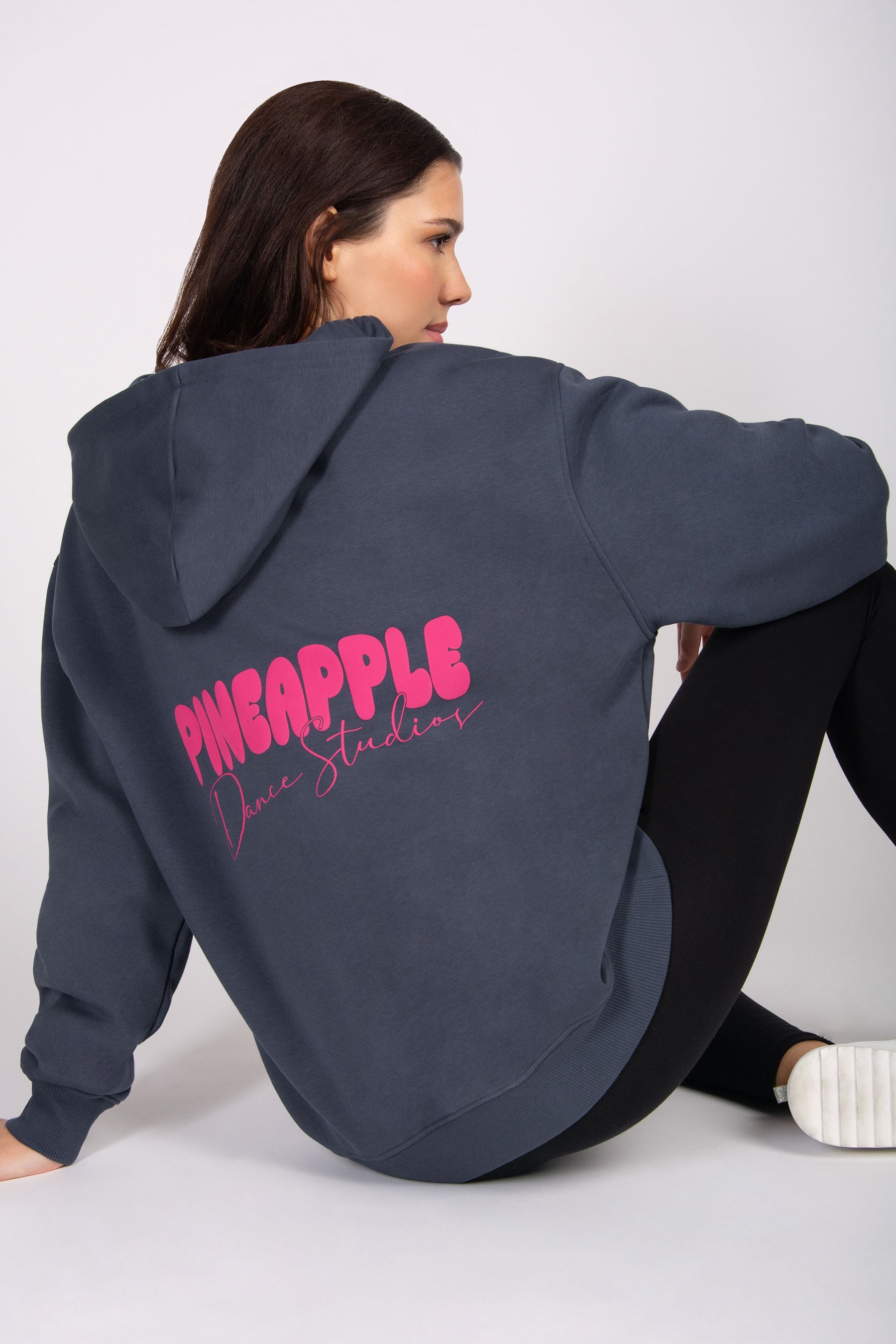 Woman wearing a dark grey hoodie with 'Pineapple Dance Studios' text on the back, sitting against a white background.