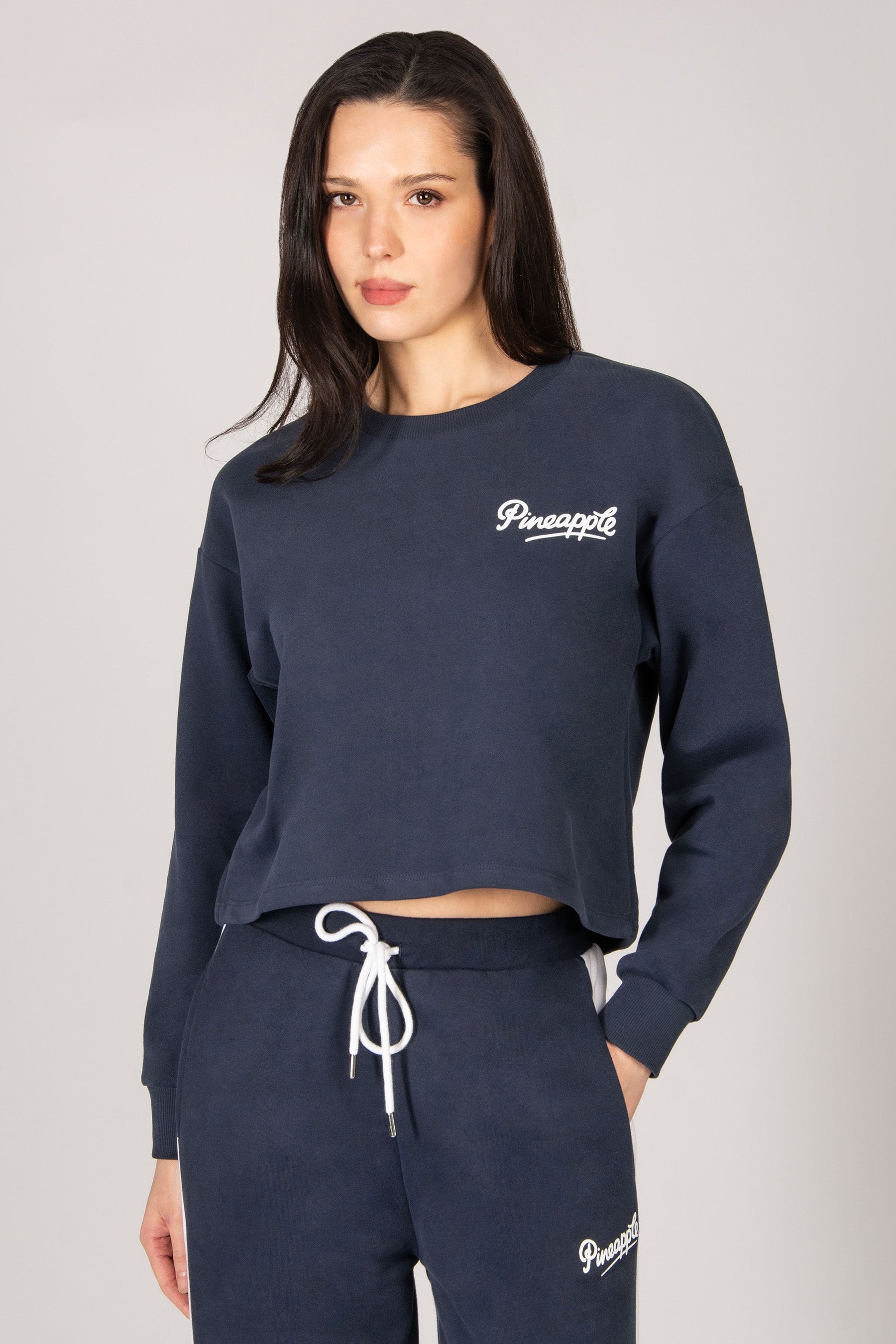 Woman wearing a dark grey sweatshirt and trousers with a brand logo on a light grey background