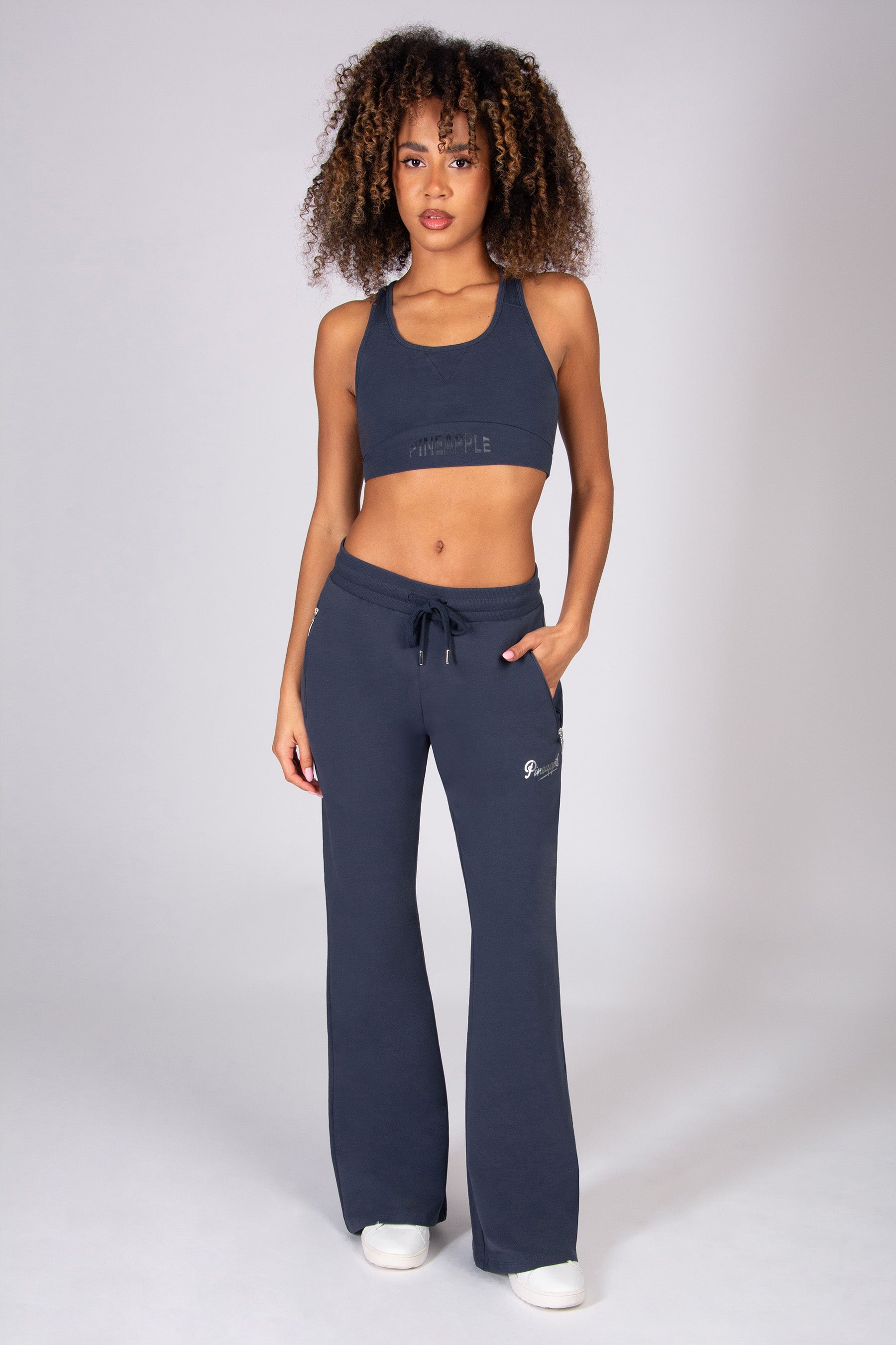 Woman wearing grey sports top and matching dark grey flared trousers with zip pockets on a light grey background. 