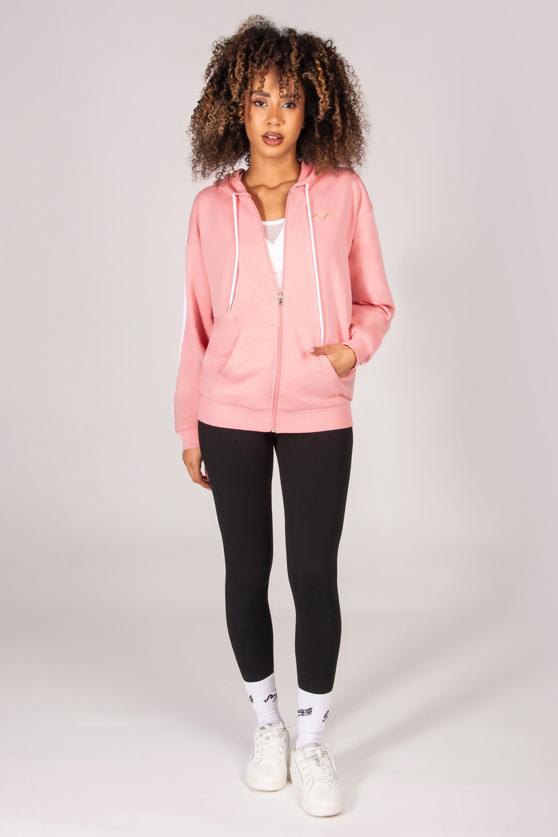 Woman wearing a light pink hoodie with zip and pockets, black leggings, and white sneakers on a light grey background