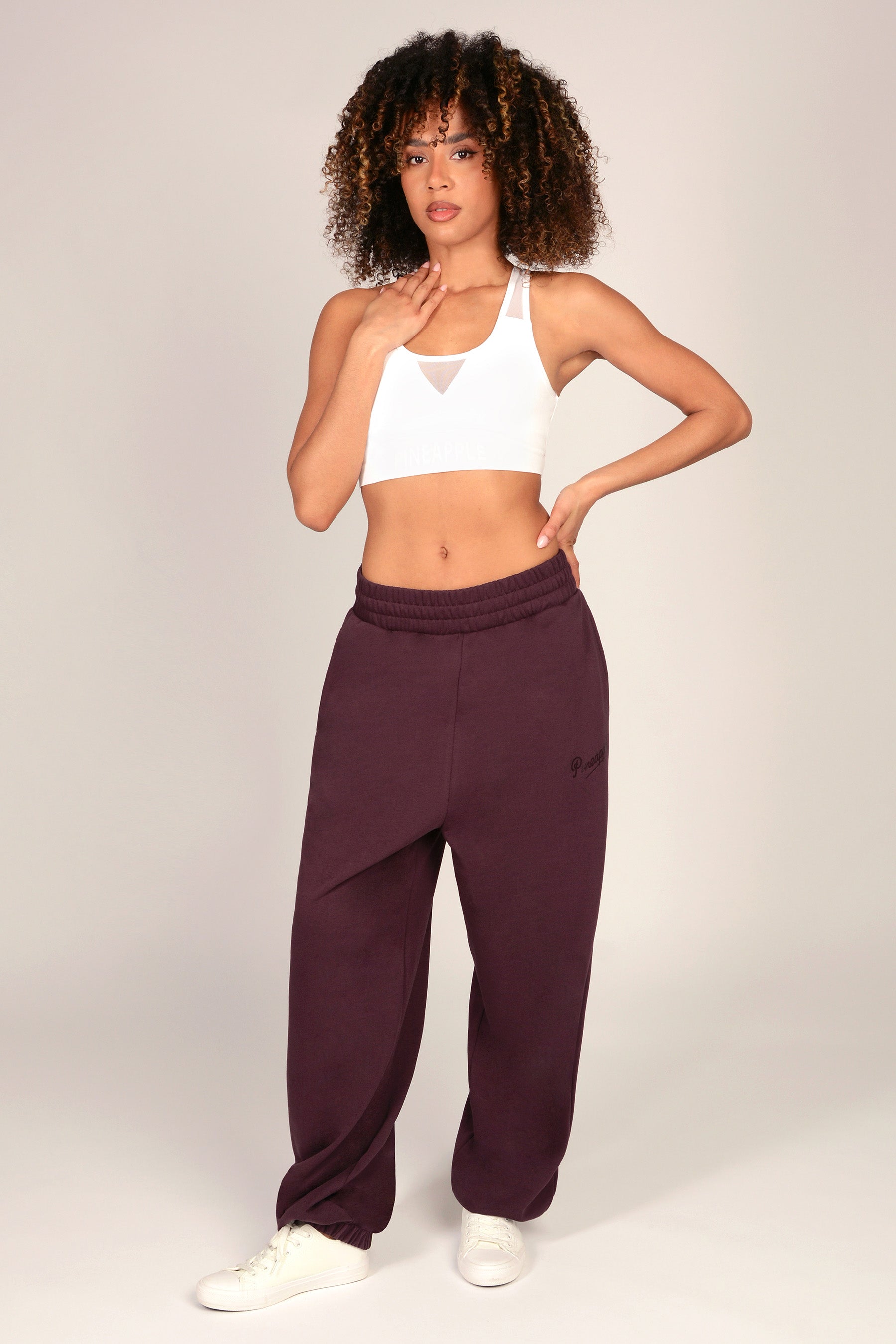 A woman modeling a pair of dark plum sweatpants with a white sports bra and white sneakers, standing against a light gray studio background.