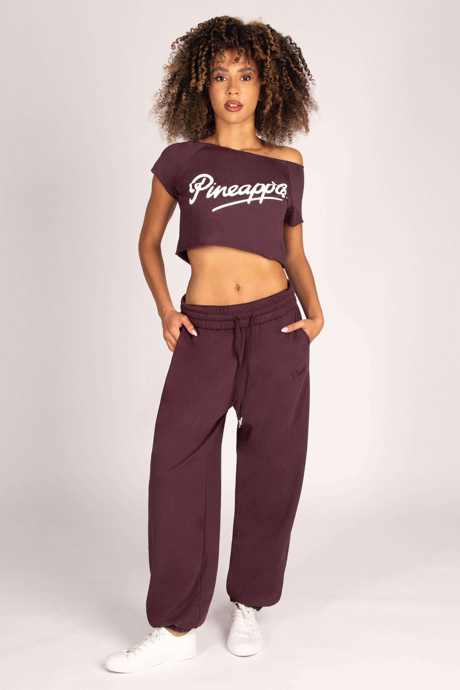 Woman wearing a brown off-shoulder crop top with matching oversized joggers against a light grey studio background.