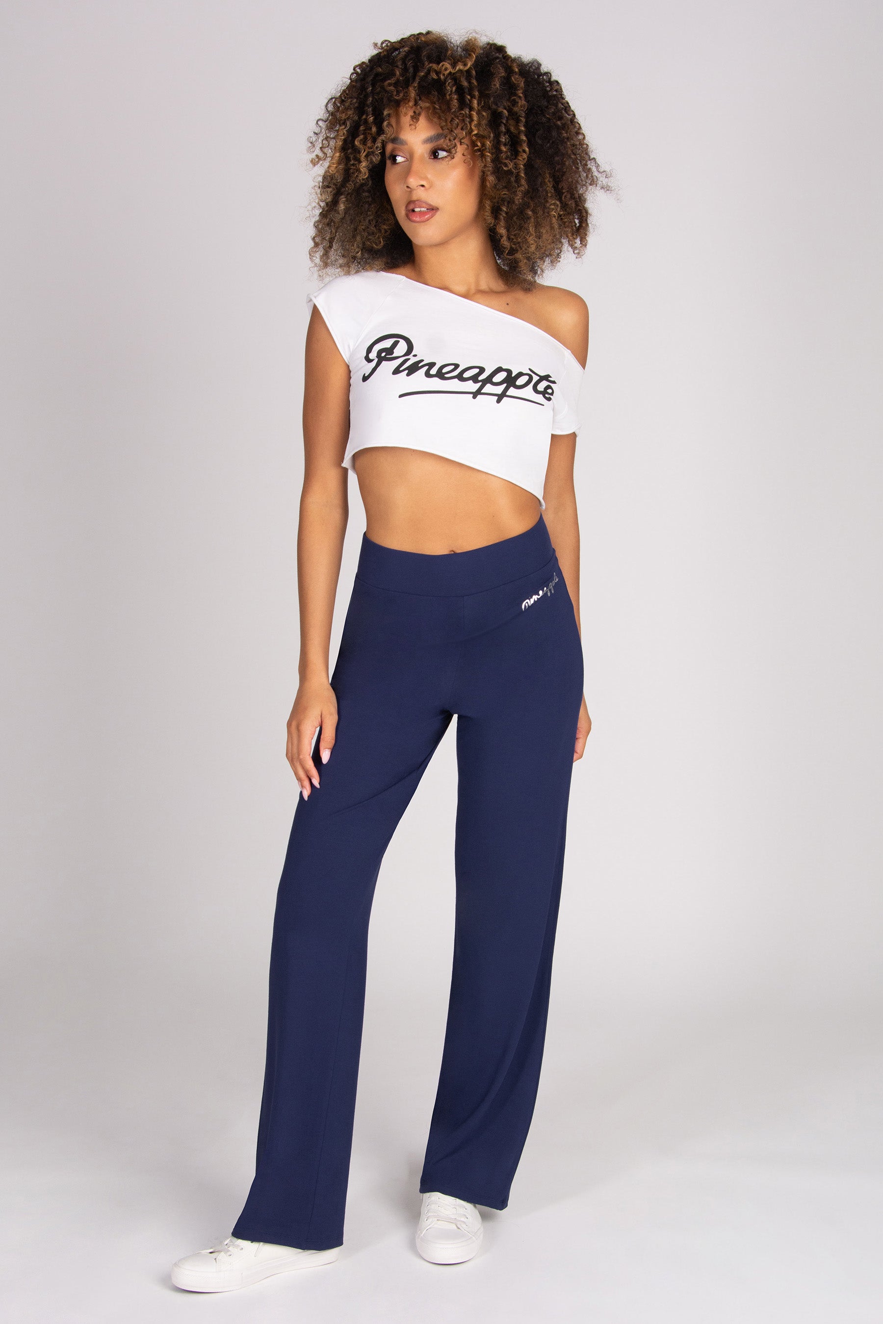 A model wearing a white off-the-shoulder crop top with 'Pineapple' printed in black script and navy blue wide-leg viscose trousers, standing against a light grey studio background.