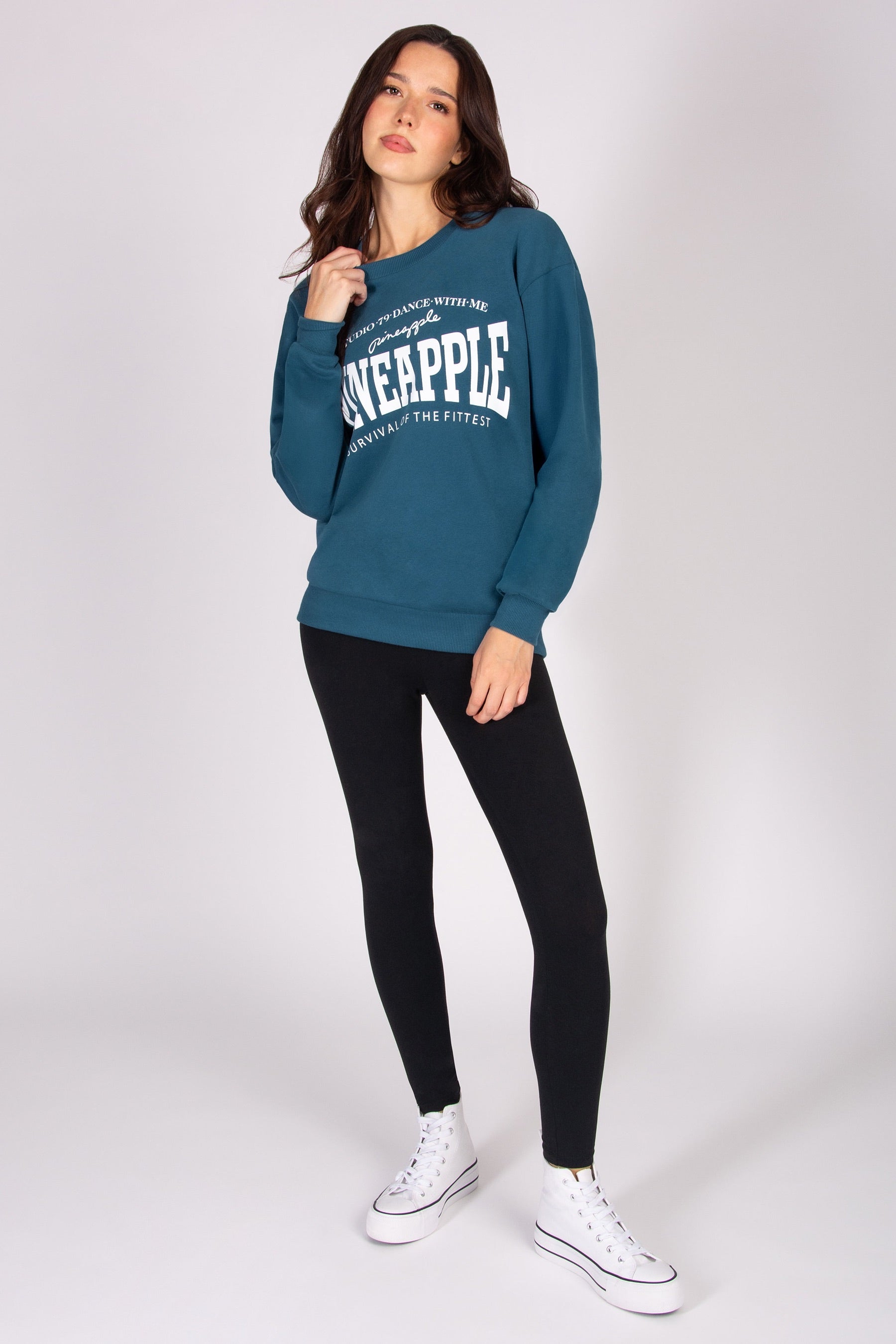 Woman wearing a teal sweatshirt with text, black leggings, and white sneakers on a white background