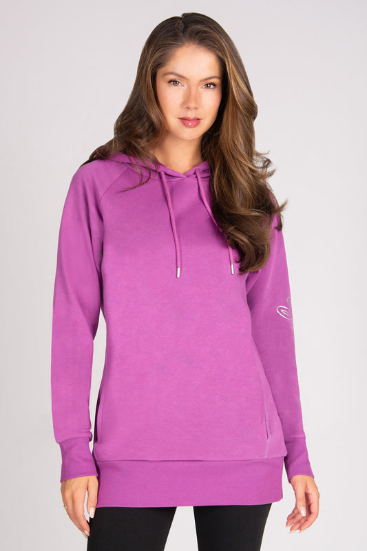 A woman modeling a bright purple long-sleeve pullover hoodie with drawstrings and a kangaroo pocket, paired with black pants, against a light gray studio background.
