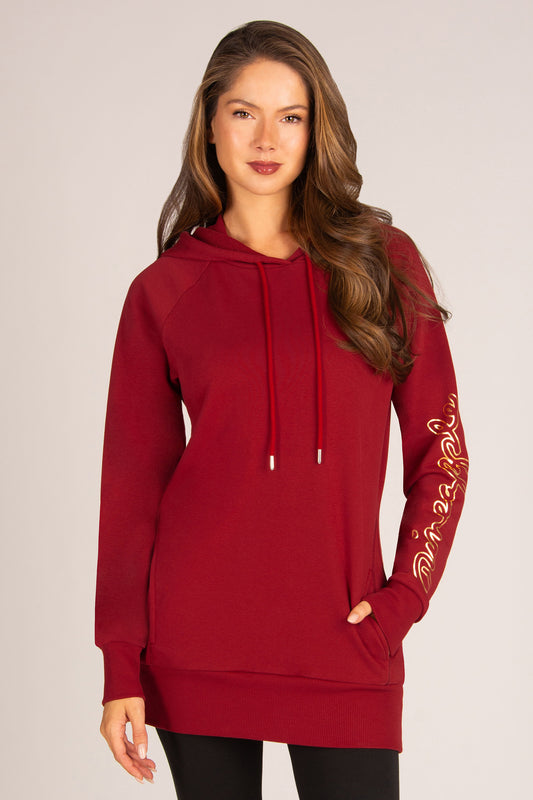 A woman modeling a long-sleeve red hoodie with gold script print on the left sleeve, set against a plain light grey studio background.