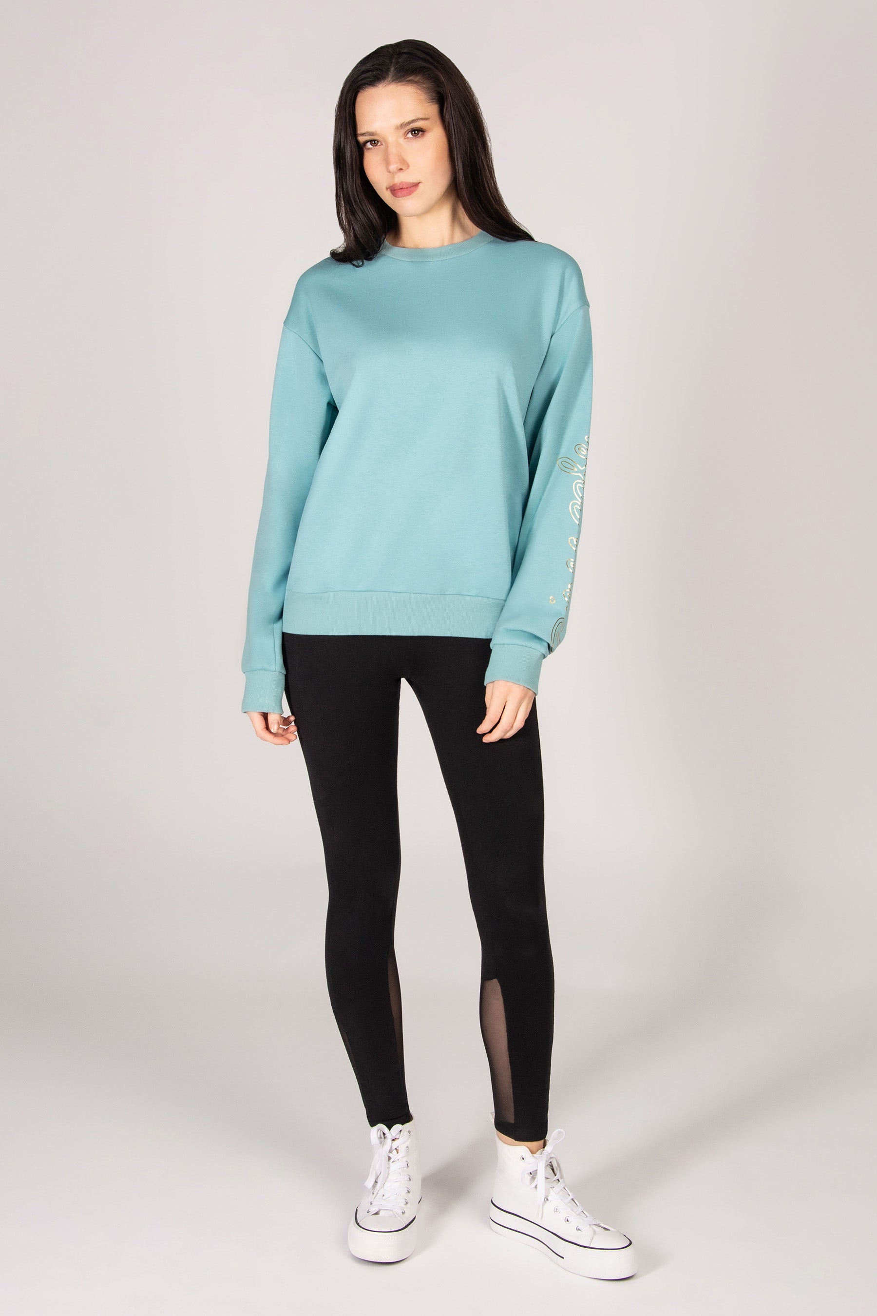 Woman wearing a turquoise oversized sweatshirt with black leggings against a grey backdrop. 