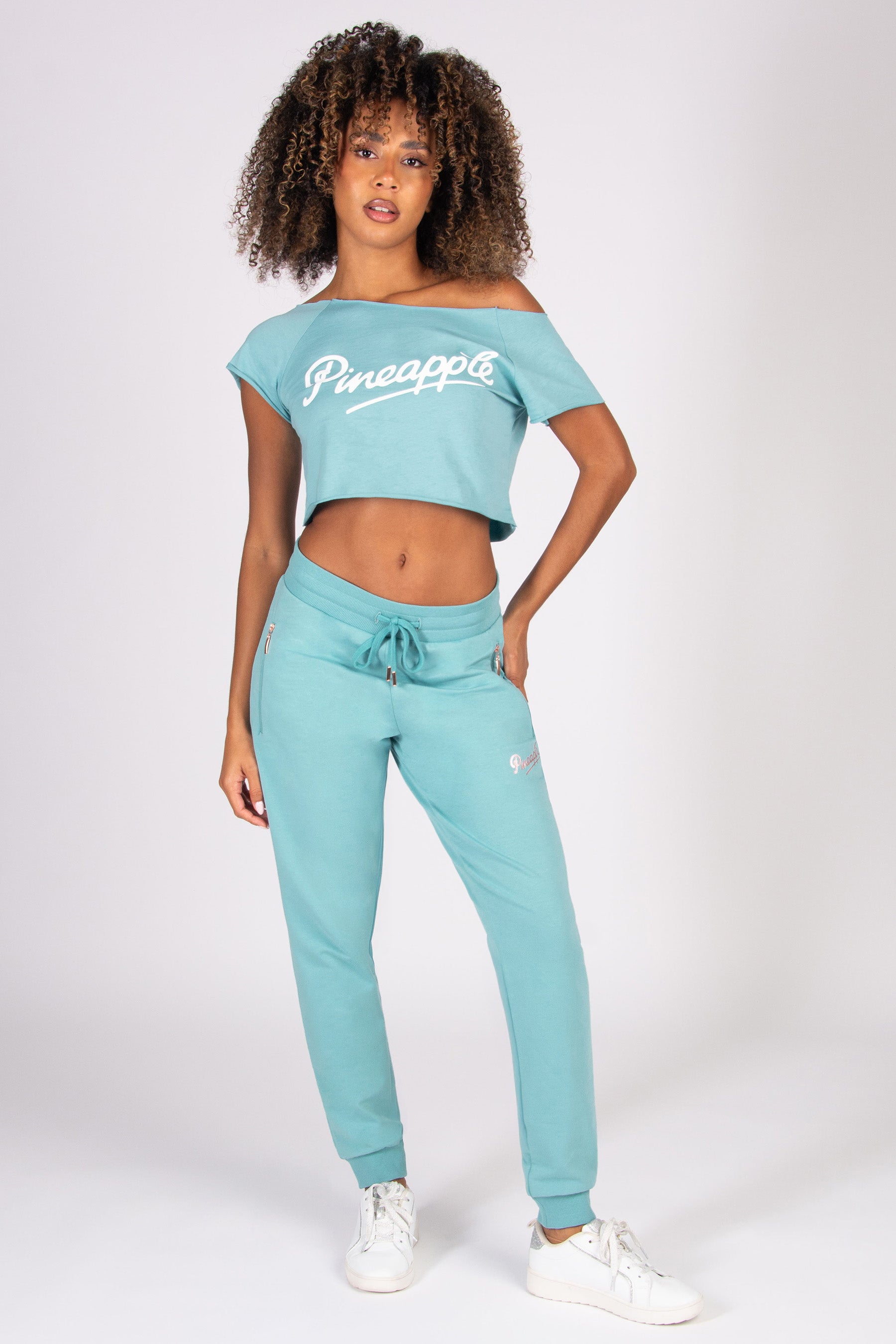 Woman wearing a teal 'Pineapple' crop top and skinny zip joggers on a white background